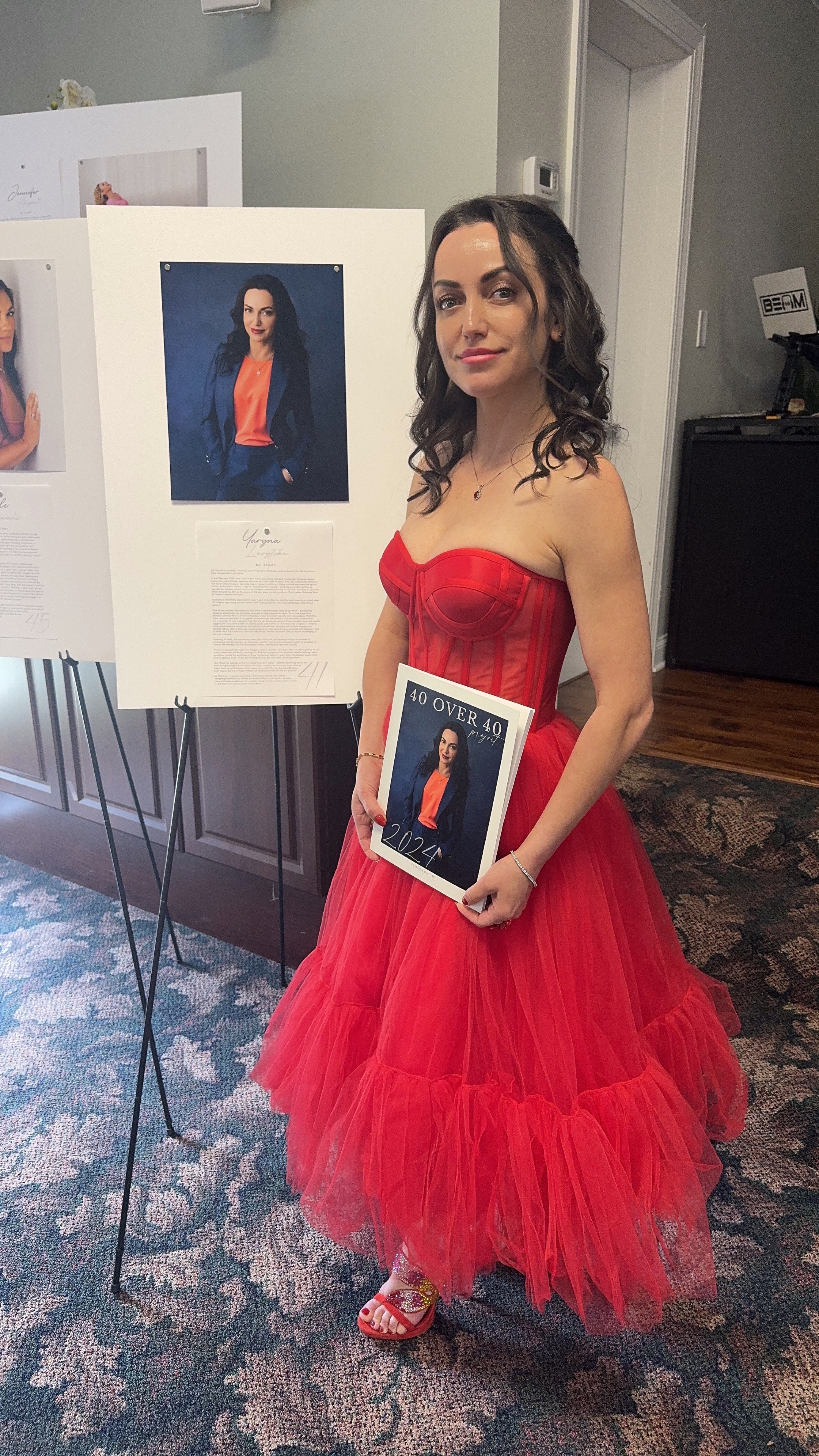 Woman in red dress holding a framed photo, standing indoors near display boards, smiling at camera.