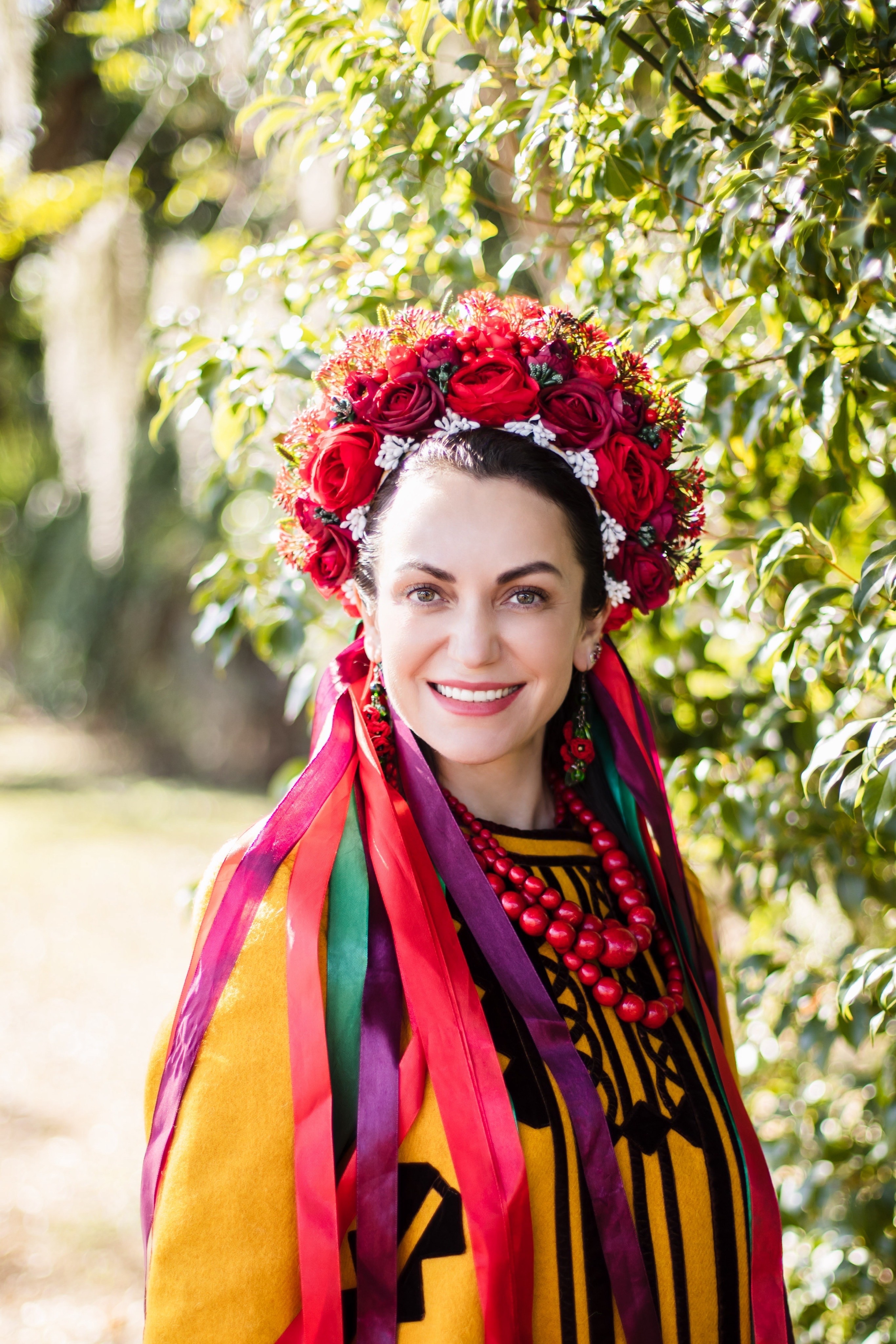 Woman wearing a colorful floral headdress and traditional clothing outdoors, smiling, surrounded by greenery.