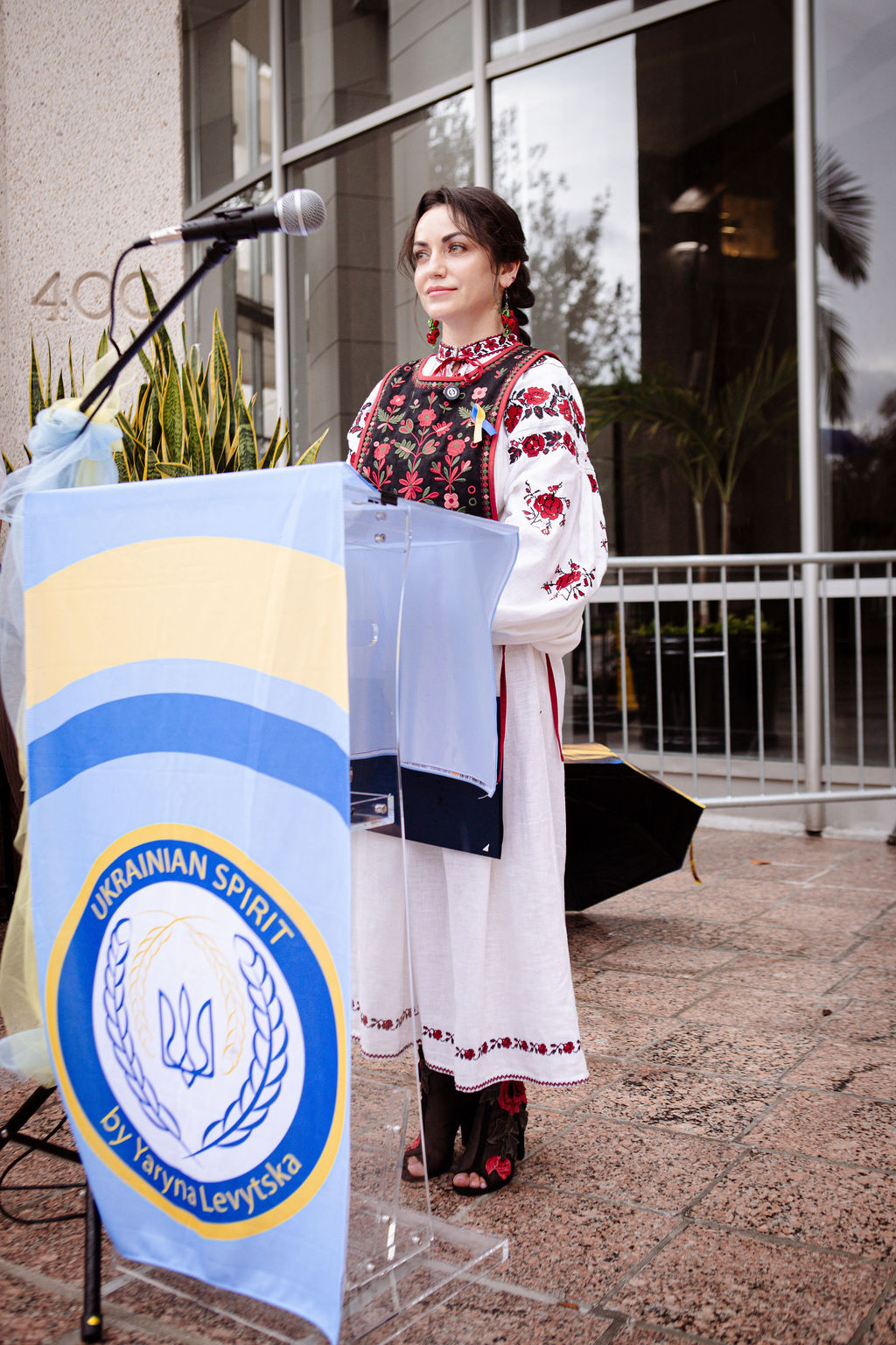 Woman in traditional dress speaking at a podium outdoors, with a building and plants in the background.