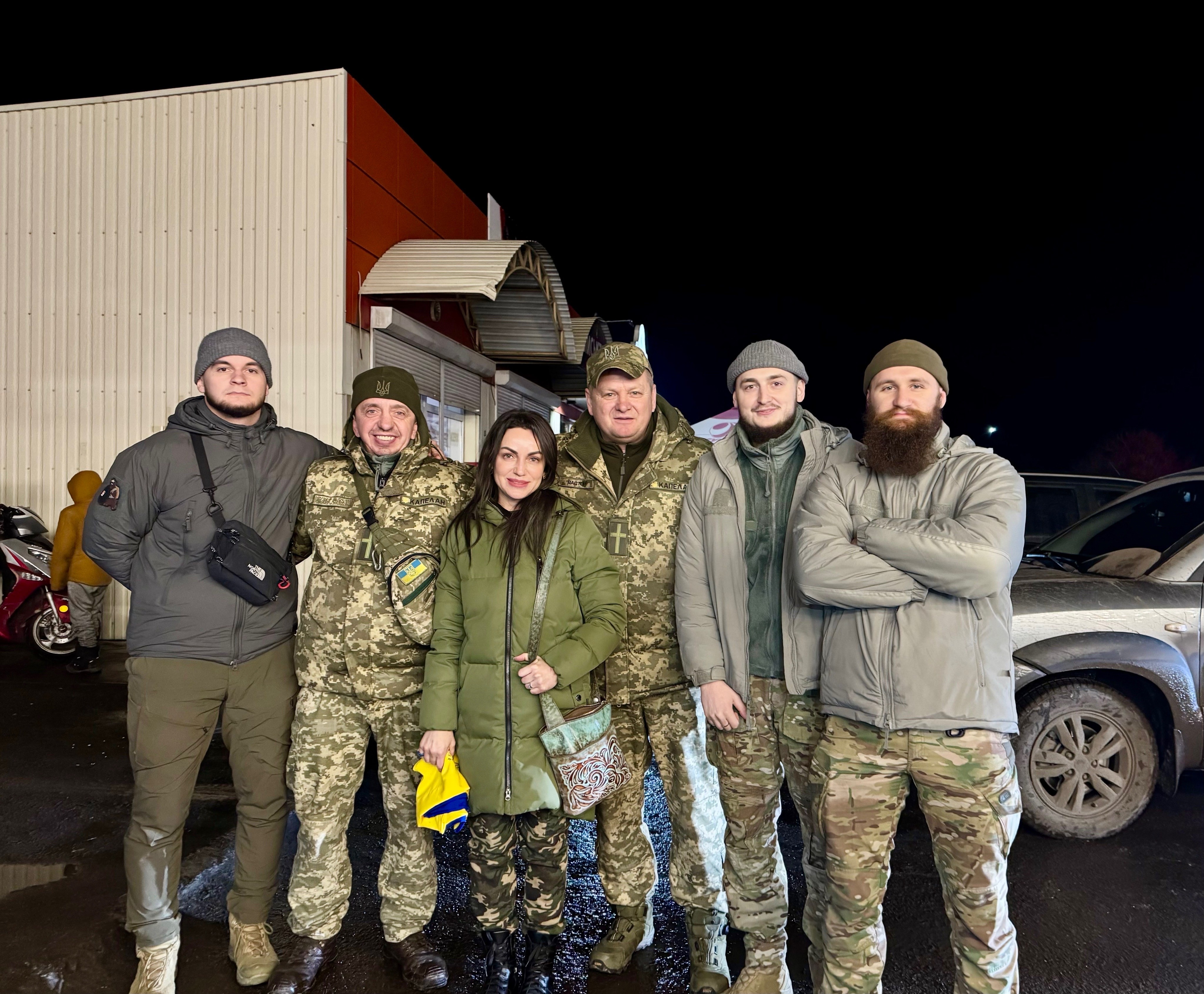 Group of six people standing outdoors at night, some in camouflage clothing, in front of vehicles and a building.