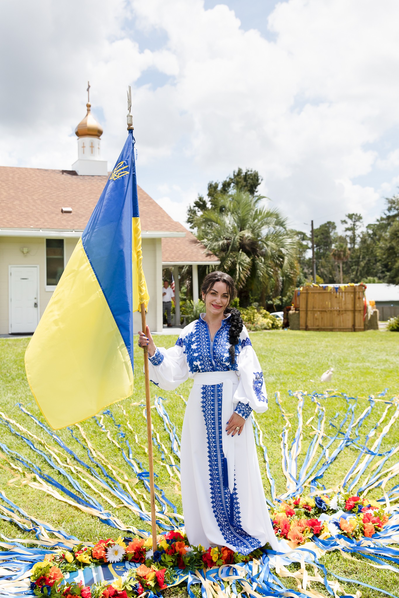Young woman in traditional dress holding a flag, standing outdoors near a church with trees and flowers around.