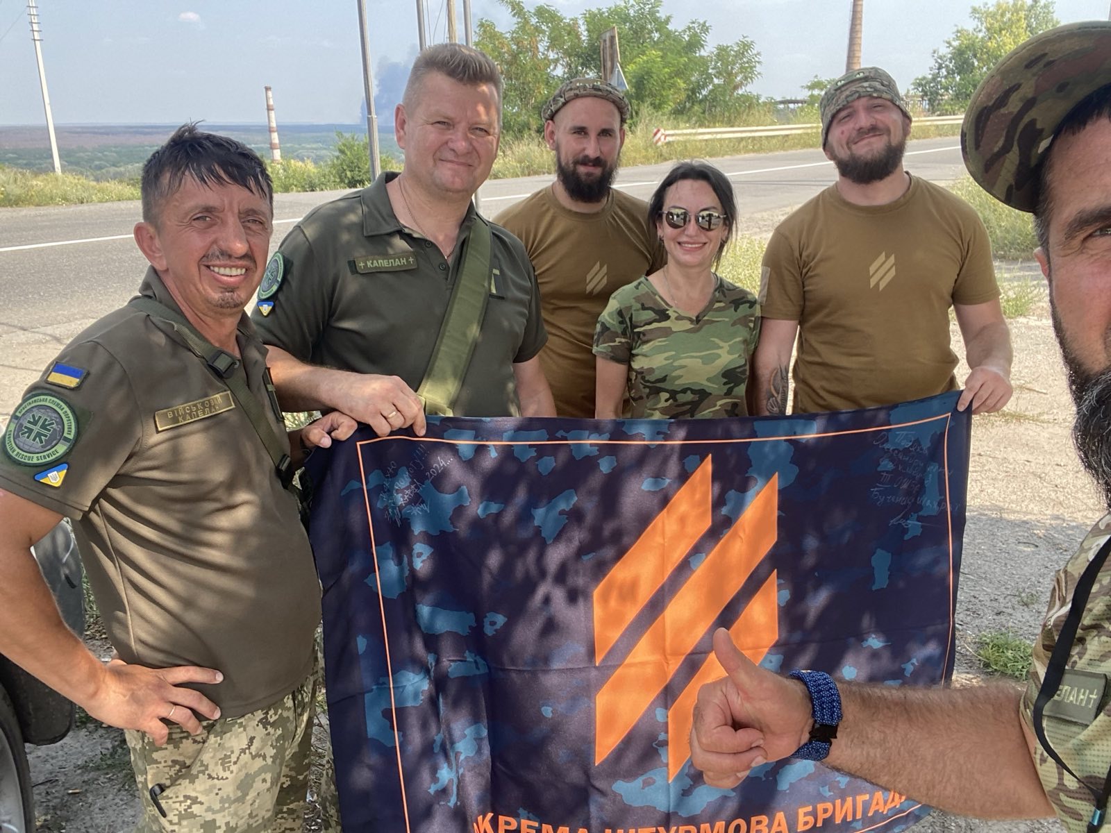 Group of six people outdoors holding a flag, some wearing military-style clothing, with trees and a road in the background.