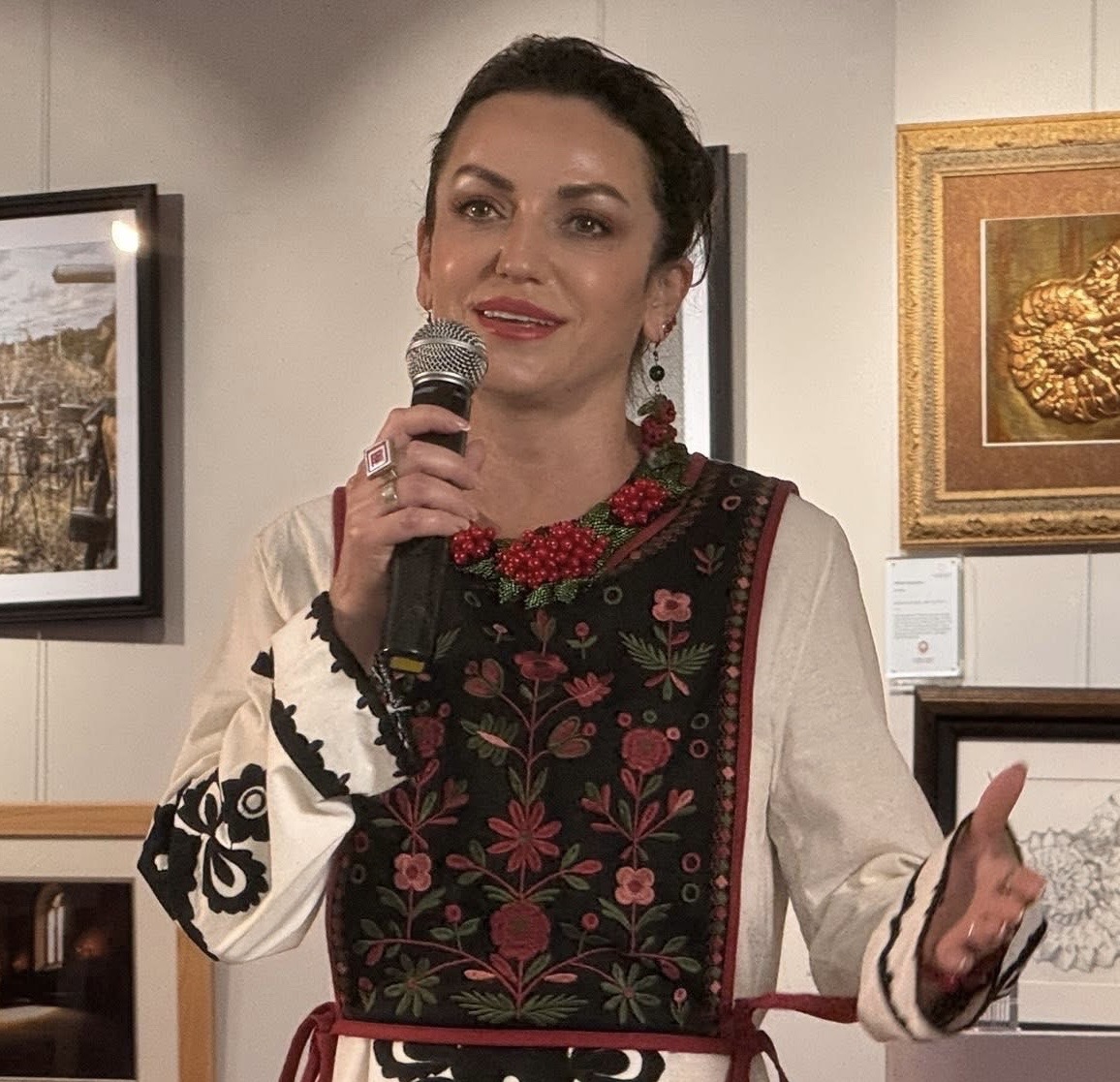 Woman speaking into a microphone, wearing traditional embroidered clothing, in an art gallery setting.