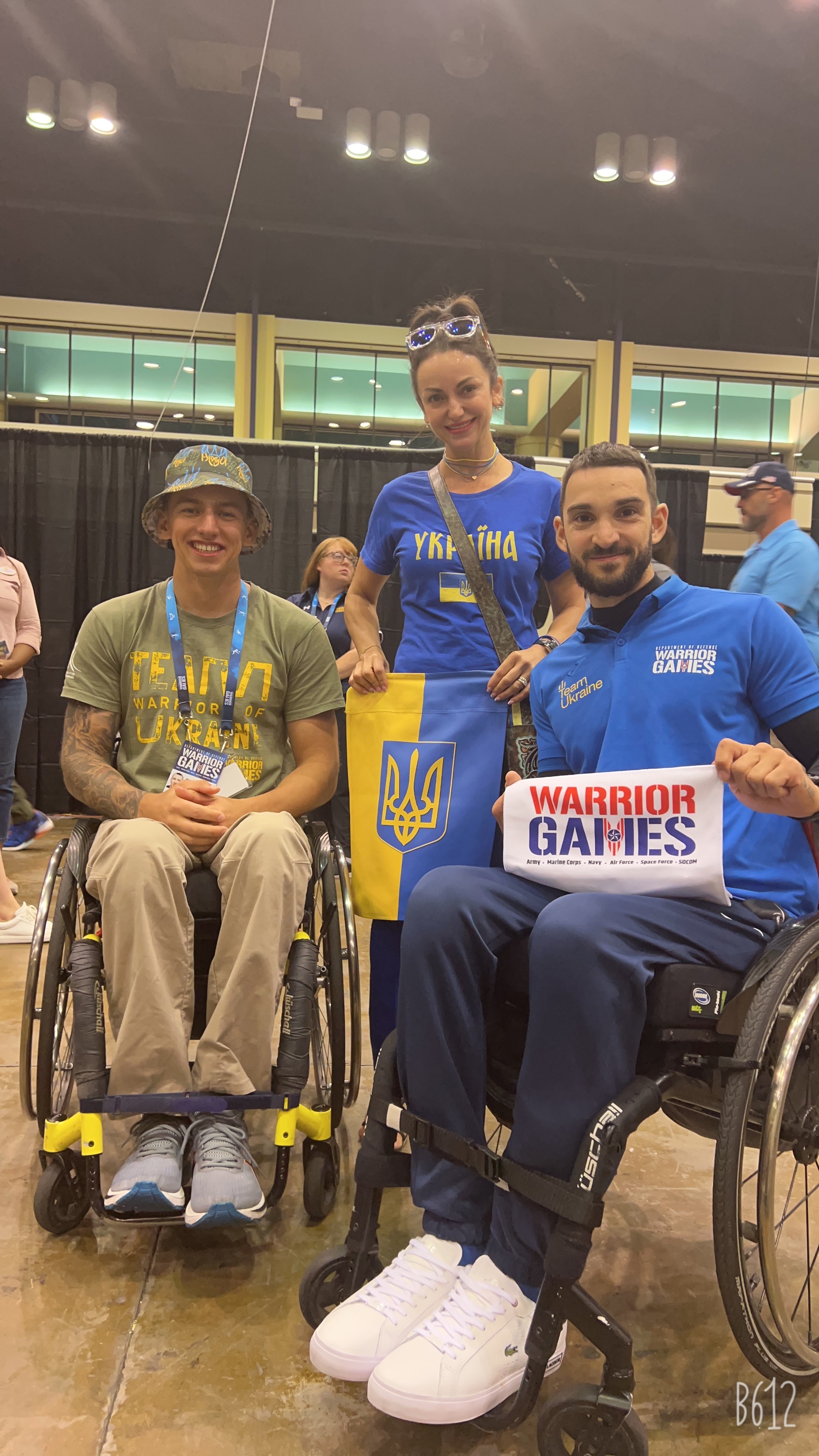 Three people, two in wheelchairs, pose indoors with medals and a flag, smiling at the camera.