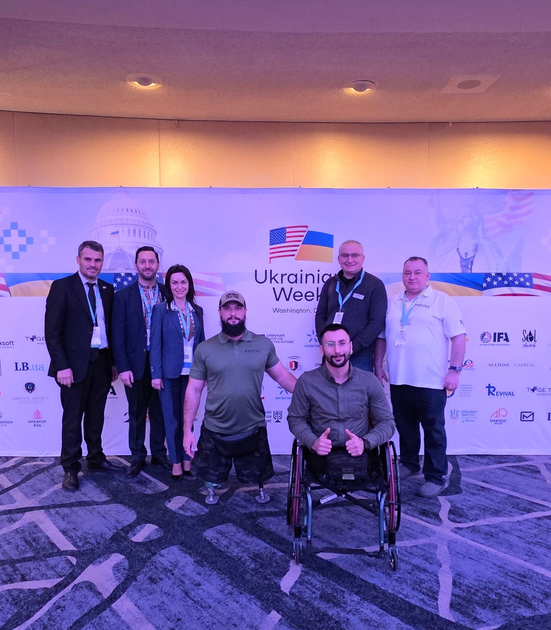 Group of seven people, including a person in a wheelchair, standing in front of a Ukraine-themed backdrop at an event.
