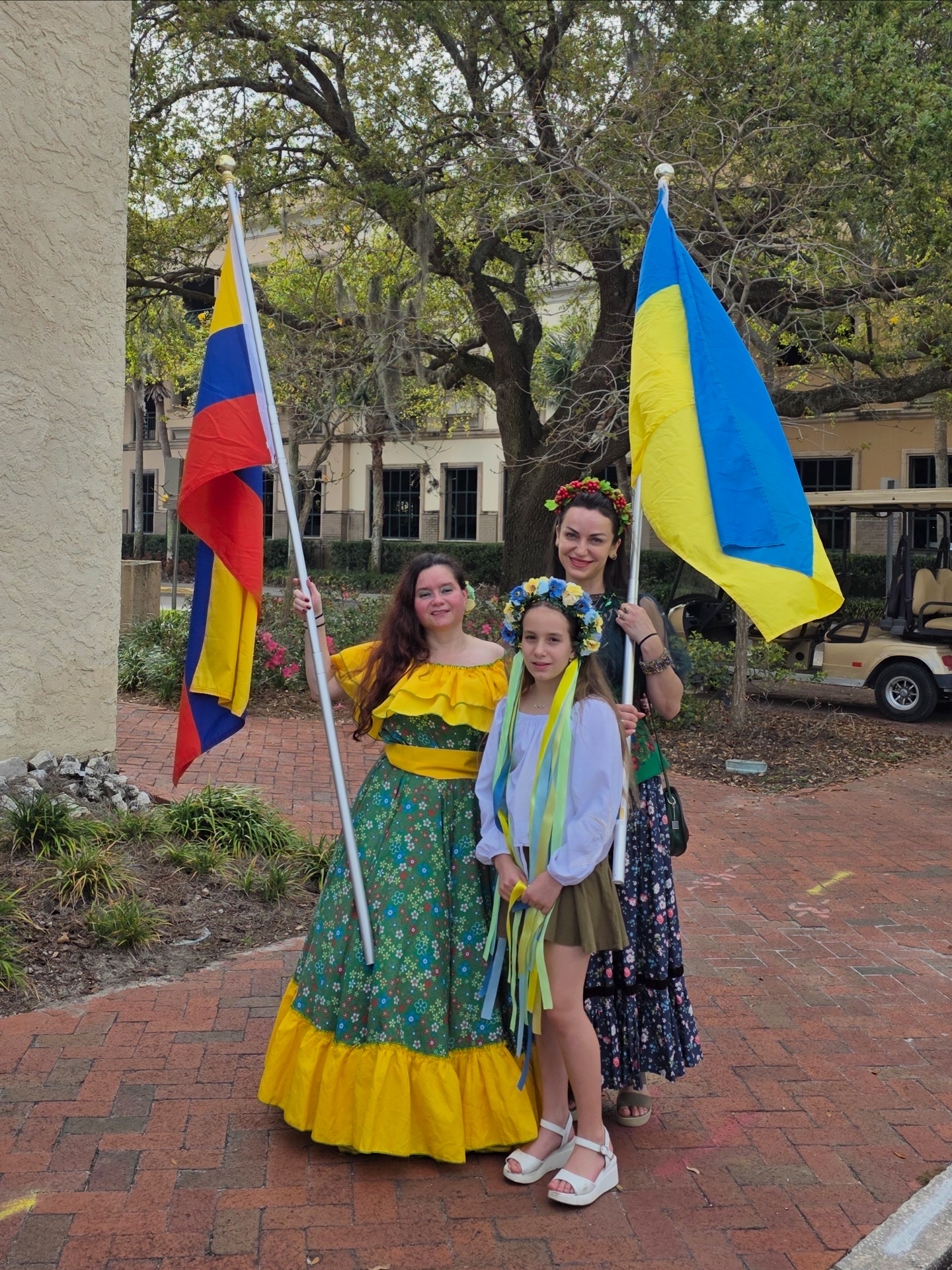 Three people standing outdoors holding flags, with trees and a building in the background.