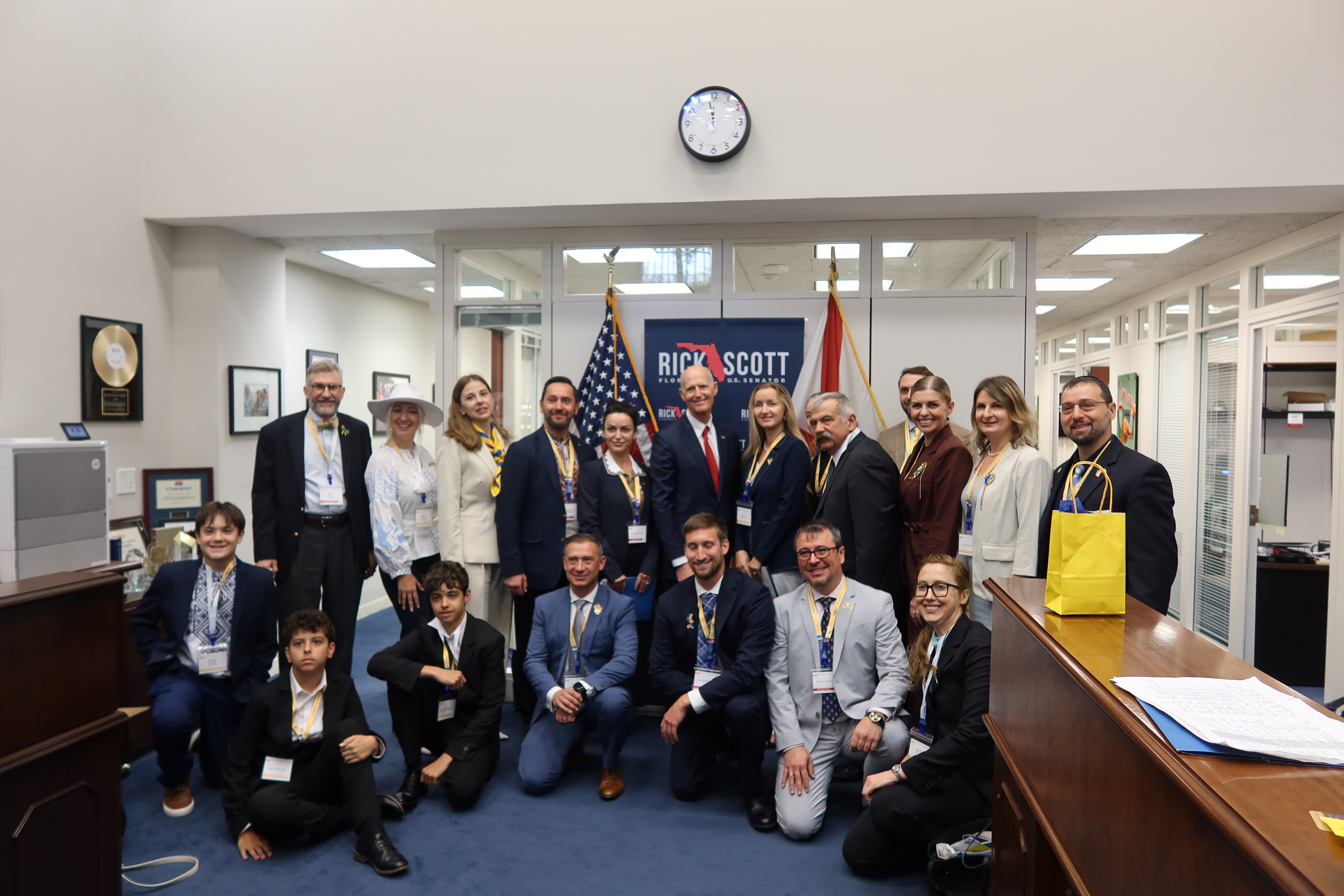 Group of people, including children and adults, gathered in an office with American flags and a clock on the wall.