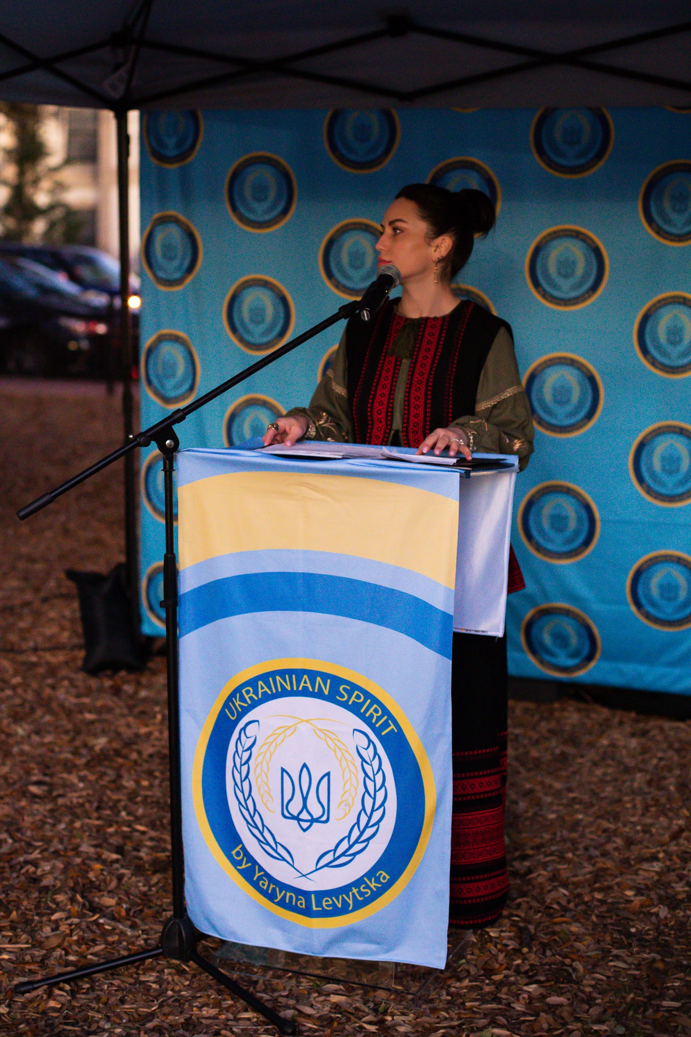 Woman standing behind a podium with a Ukrainian flag, outdoors with a blue patterned backdrop.