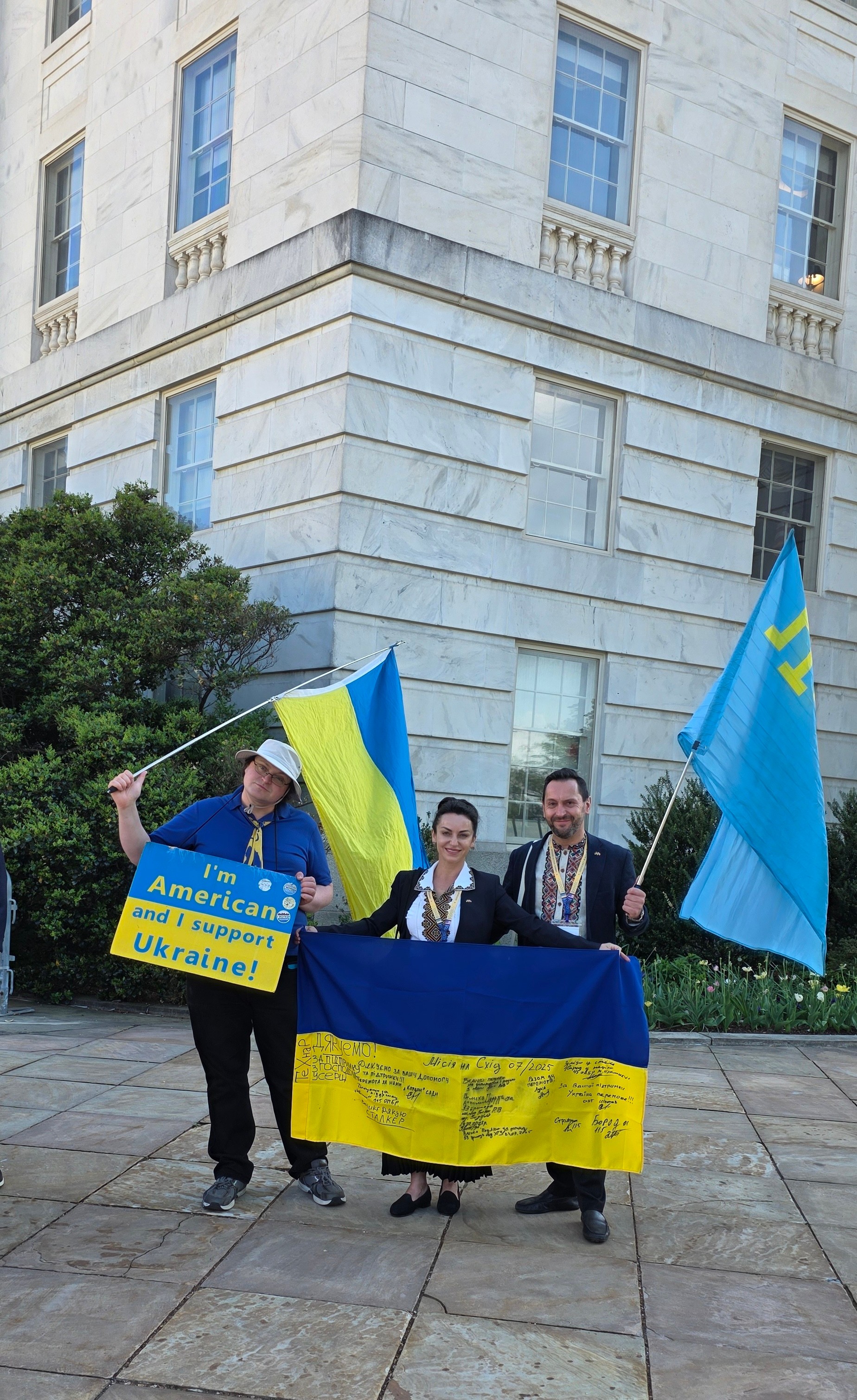 Three people holding Ukrainian flags and a yellow banner in front of a large stone building, with greenery nearby.