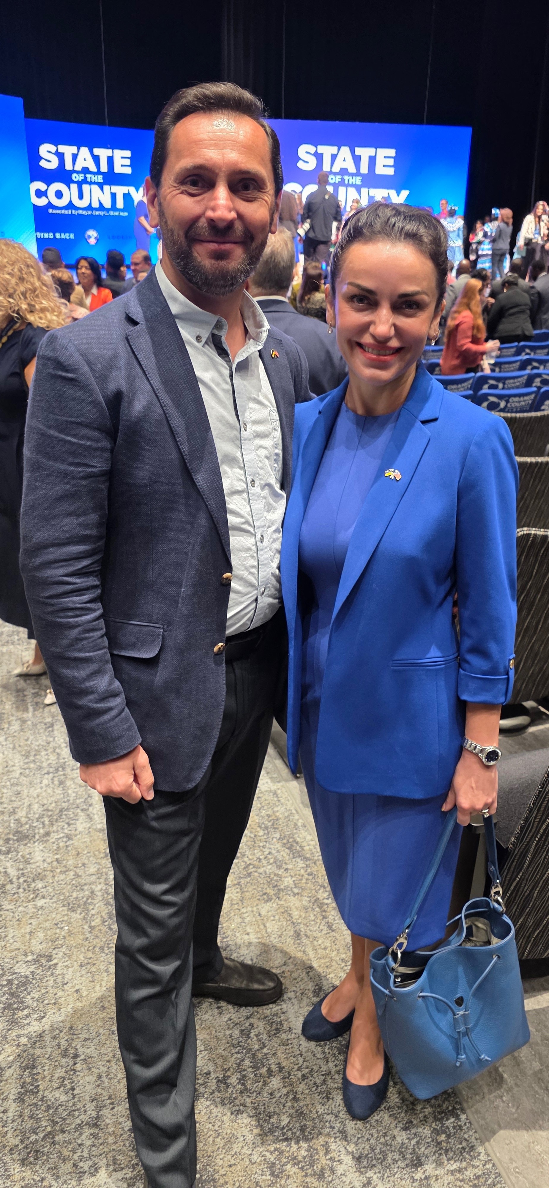 Man and woman in blue suits standing together at an event, with a large screen in the background displaying text.