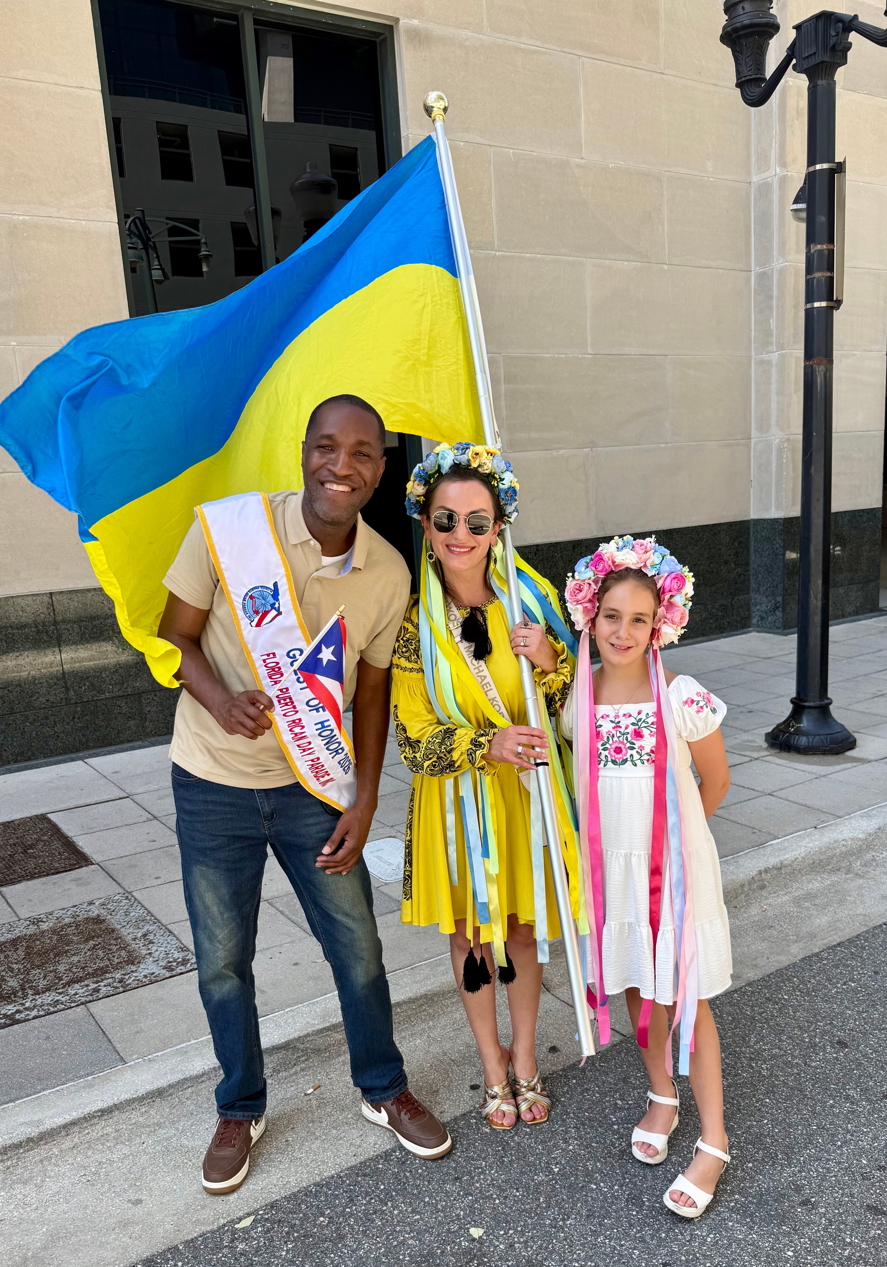Three people standing outdoors, holding a Ukrainian flag, smiling, with a building and streetlamp in the background.