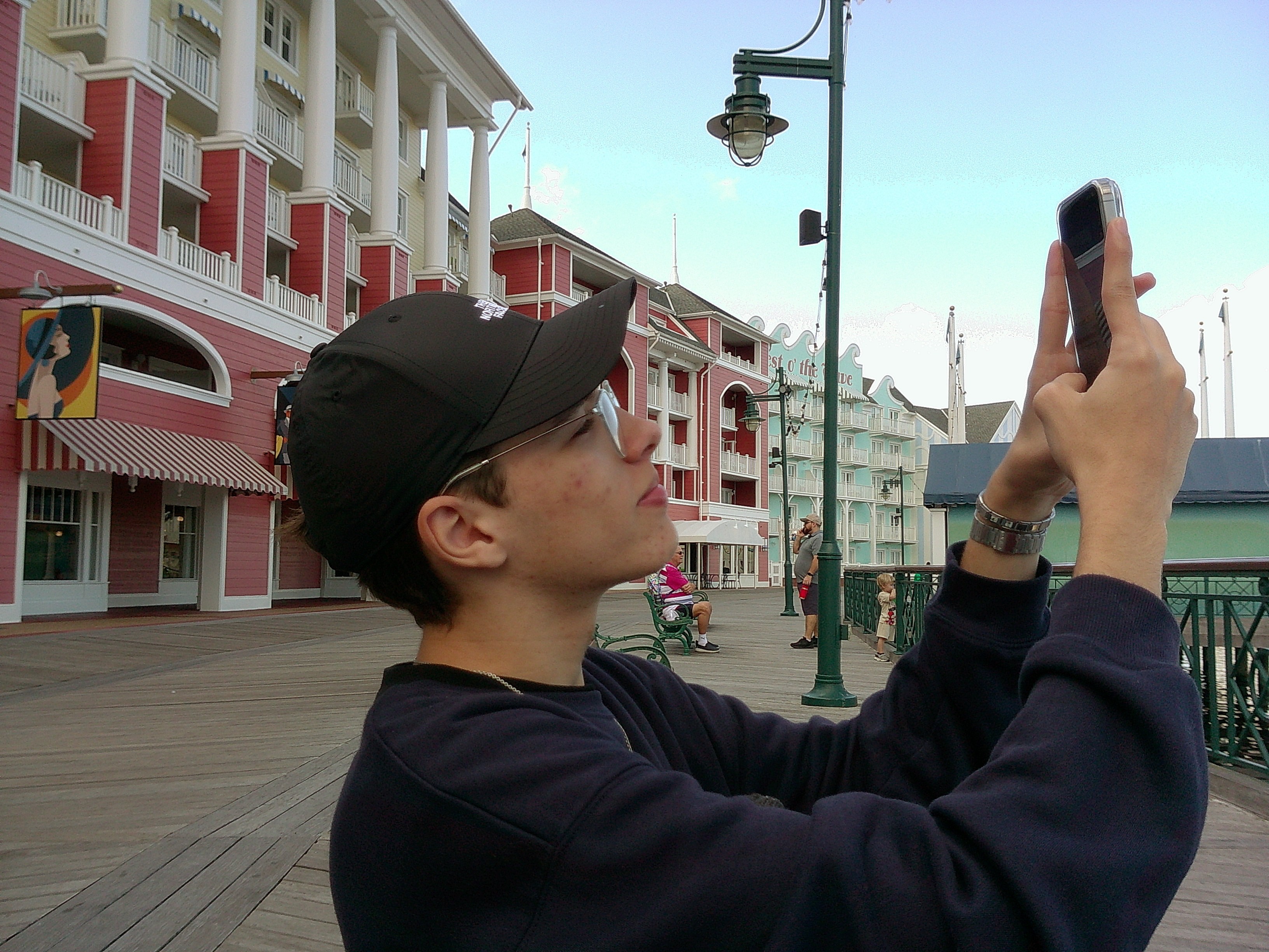 Person wearing a black cap and dark clothing taking a photo with a smartphone on a boardwalk, colorful buildings in background.