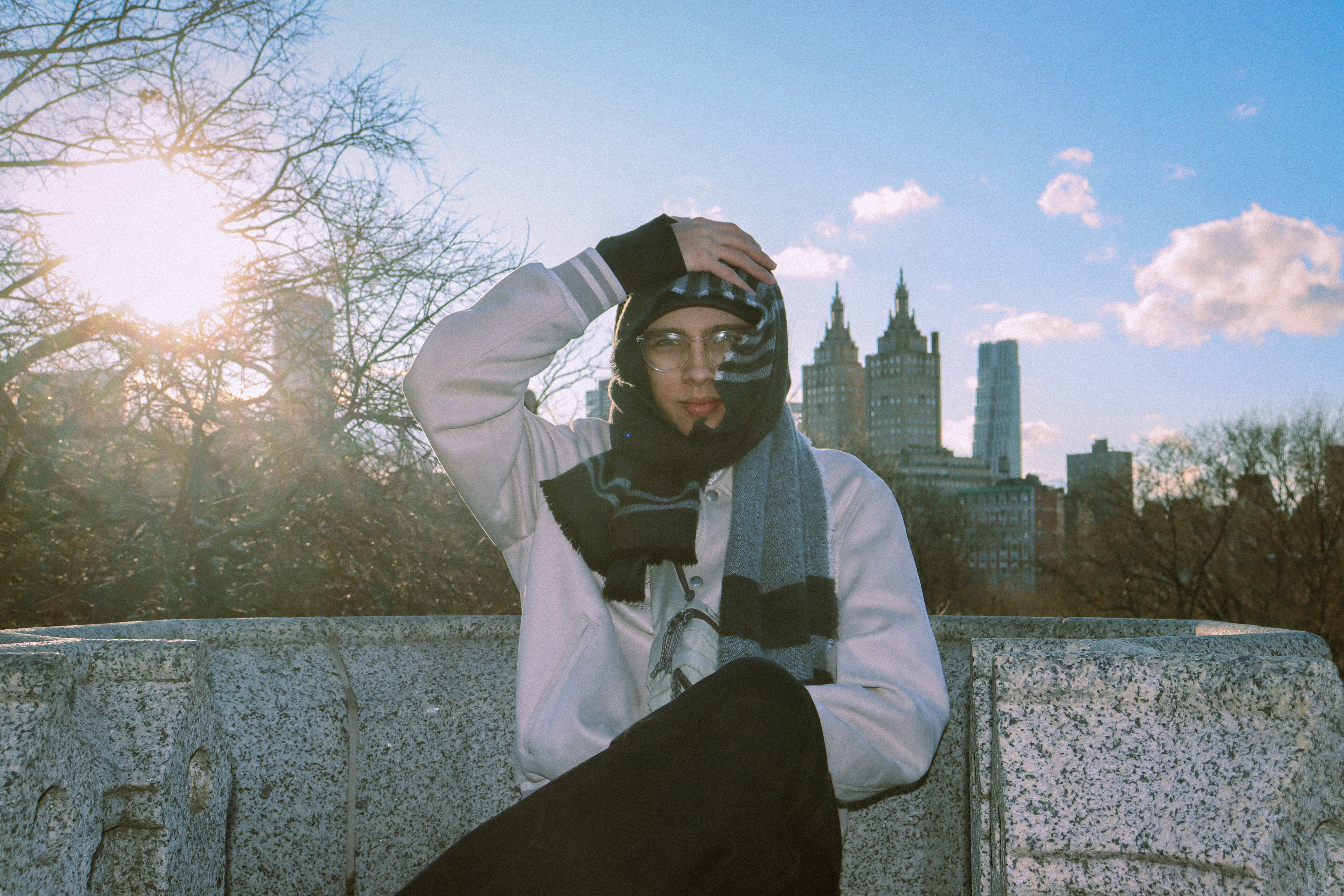 Person wearing glasses, scarf, and jacket standing outdoors with city skyline and trees in background.