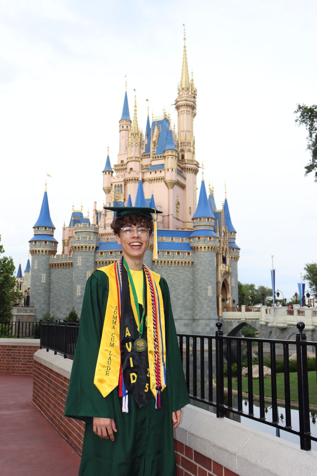 Person in graduation gown and cap standing in front of a castle-like structure with towers and spires.