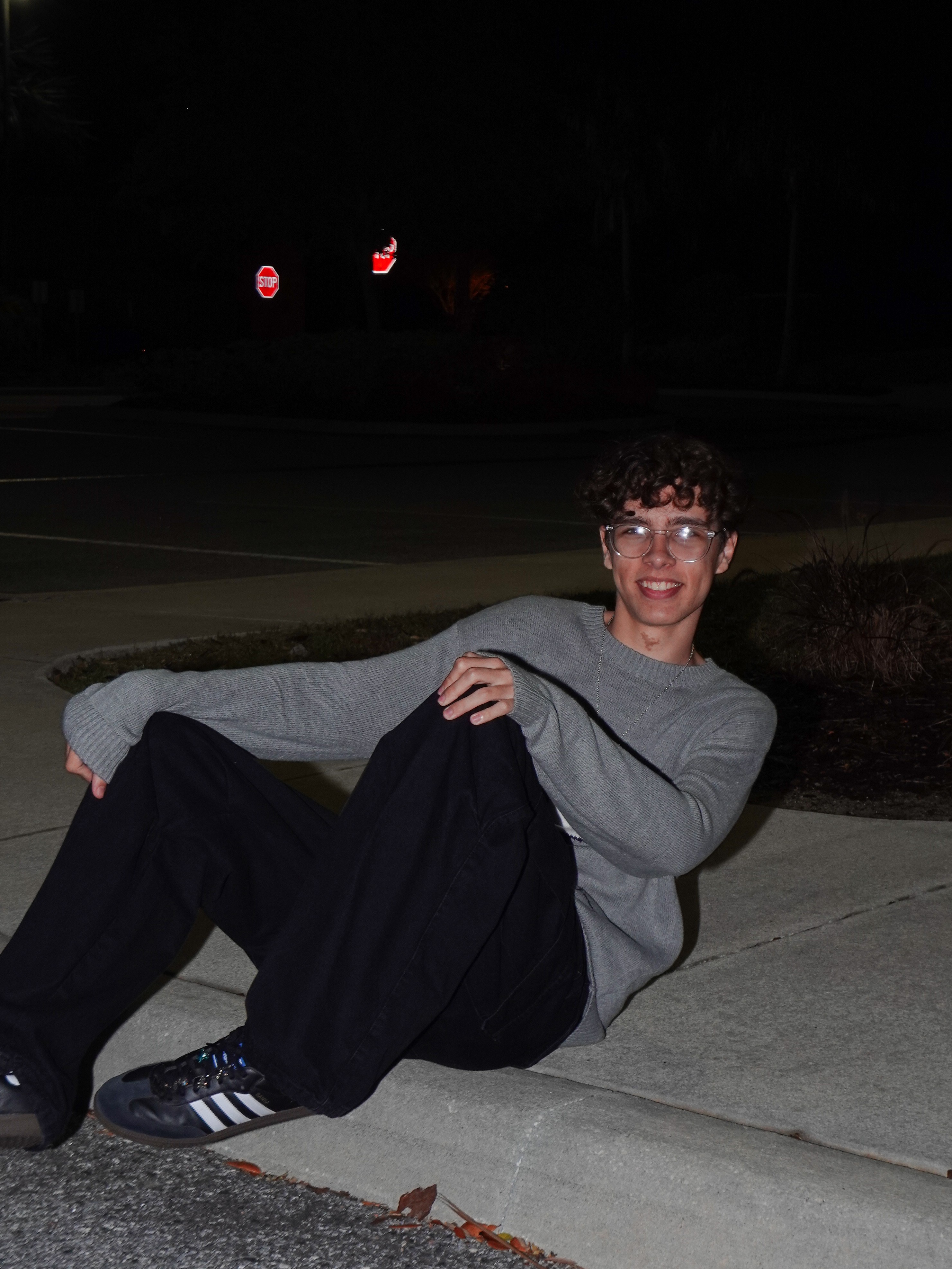 Person sitting on sidewalk at night, smiling, wearing glasses, gray long-sleeve shirt, black pants, and sneakers.