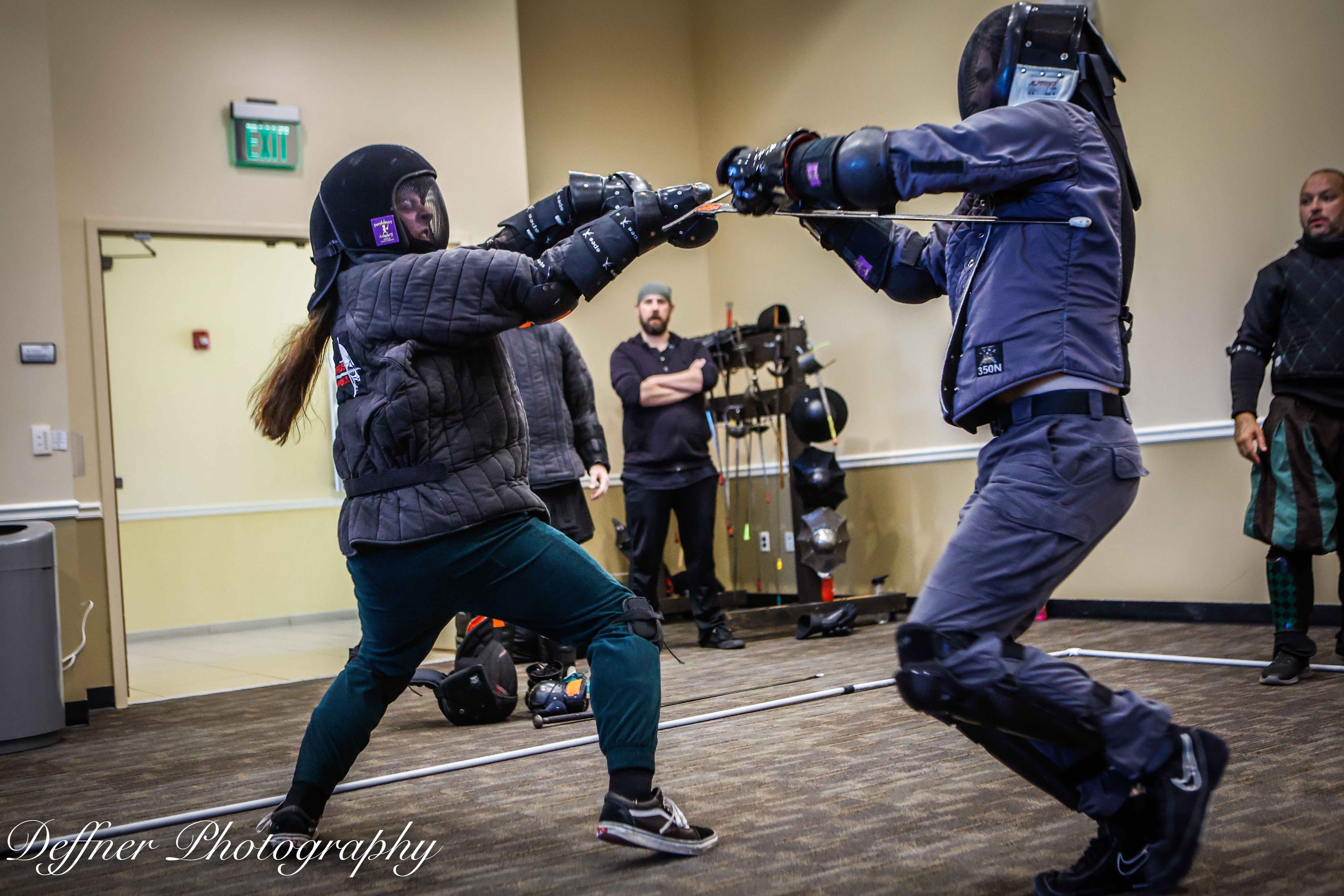 Two people in protective gear sparring with padded weapons in a room, others watching in the background.