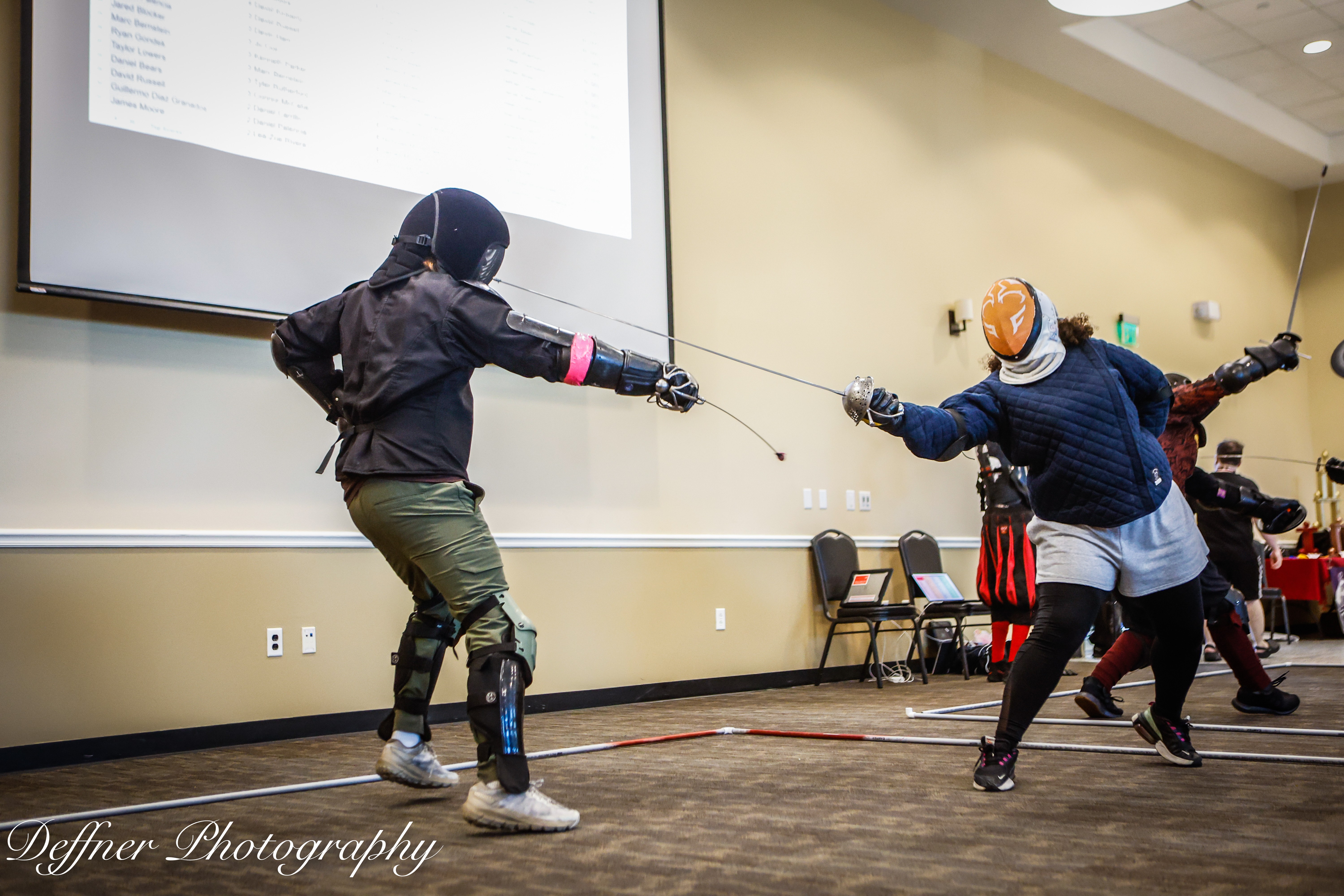 Two people engaged in a fencing match indoors, with a large screen and chairs in the background.
