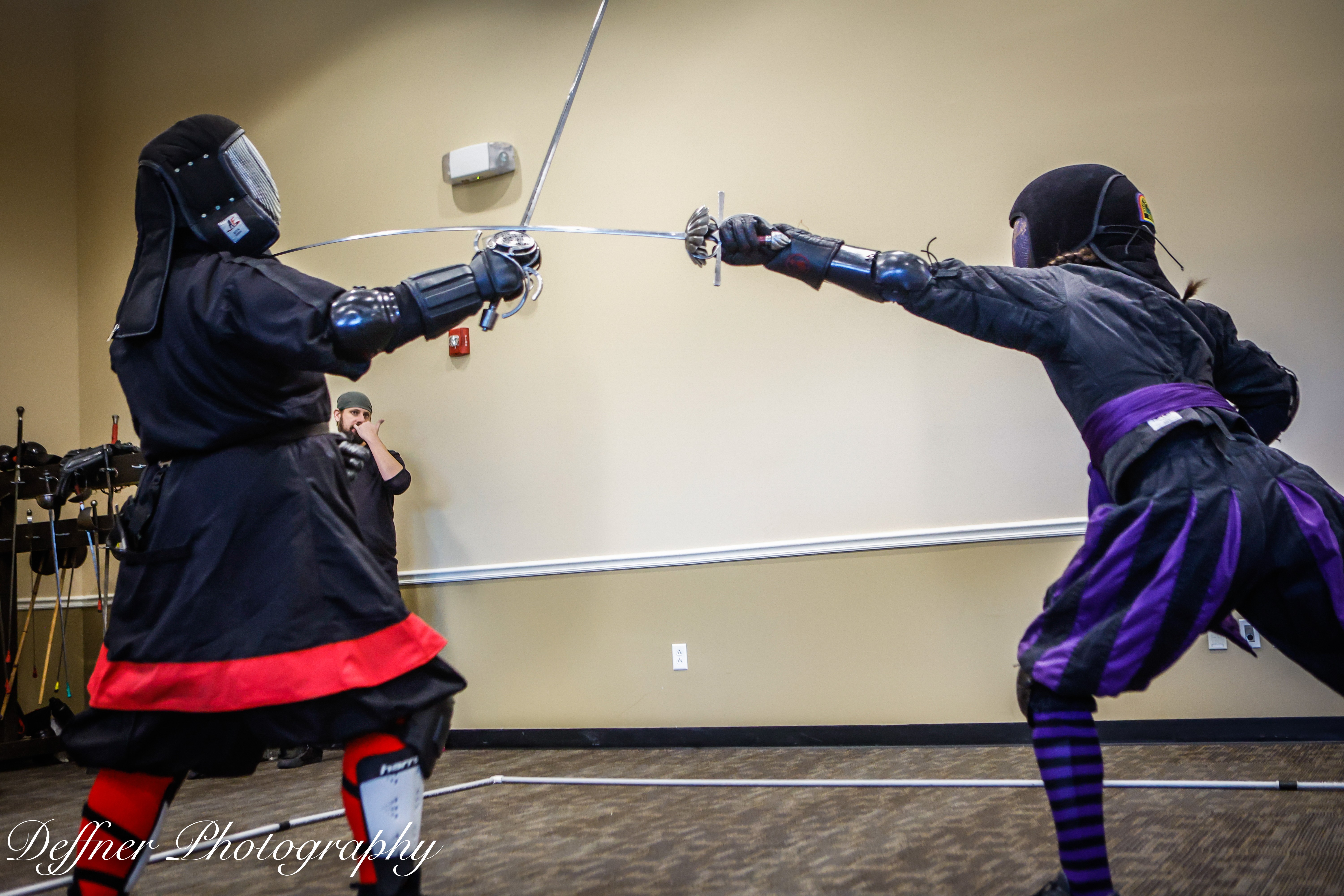 Two people in fencing gear sparring with swords in a room with beige walls and a wooden floor.