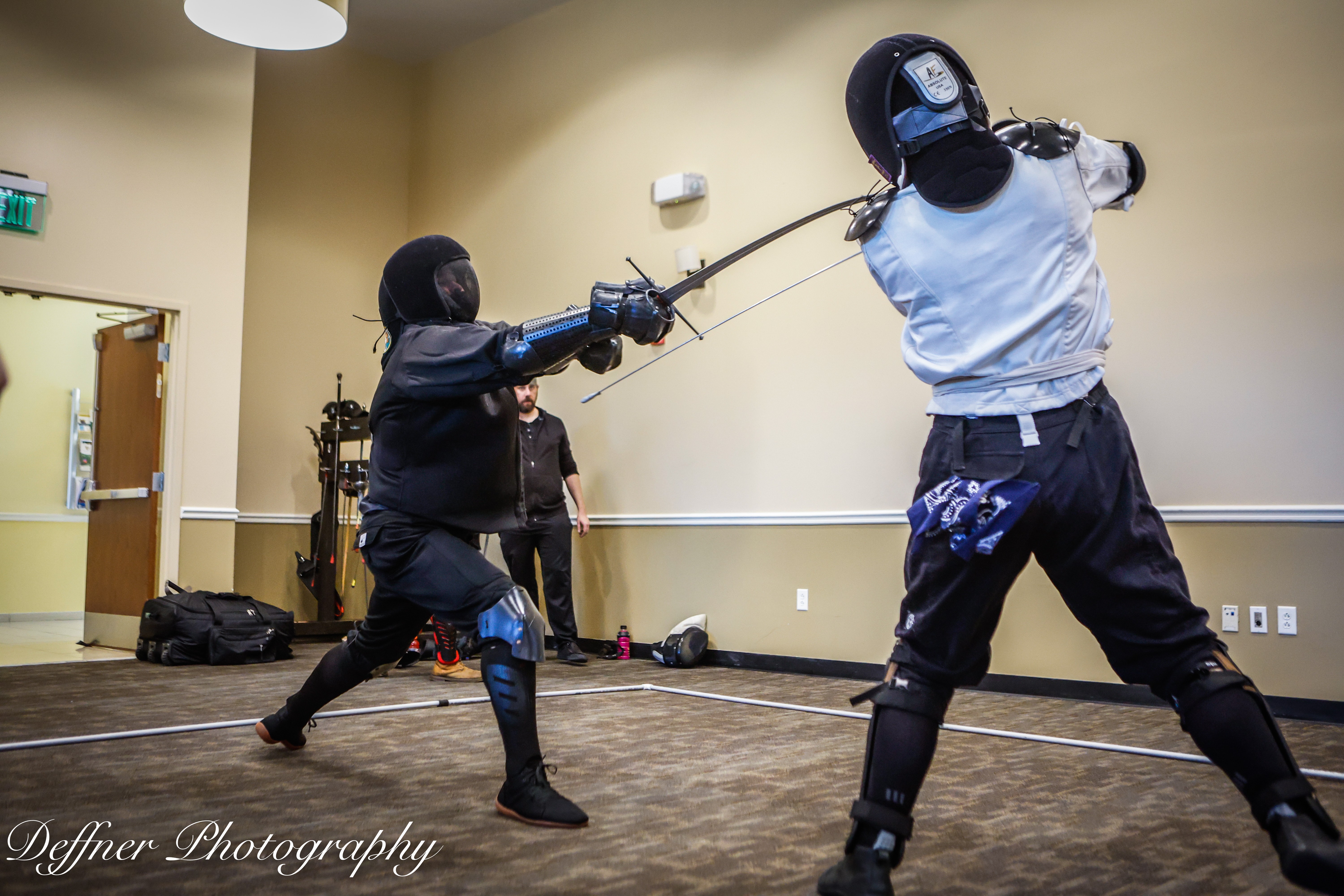 Two people engaged in a fencing match with protective gear indoors, one person lunging and the other blocking, in a room with beige walls.
