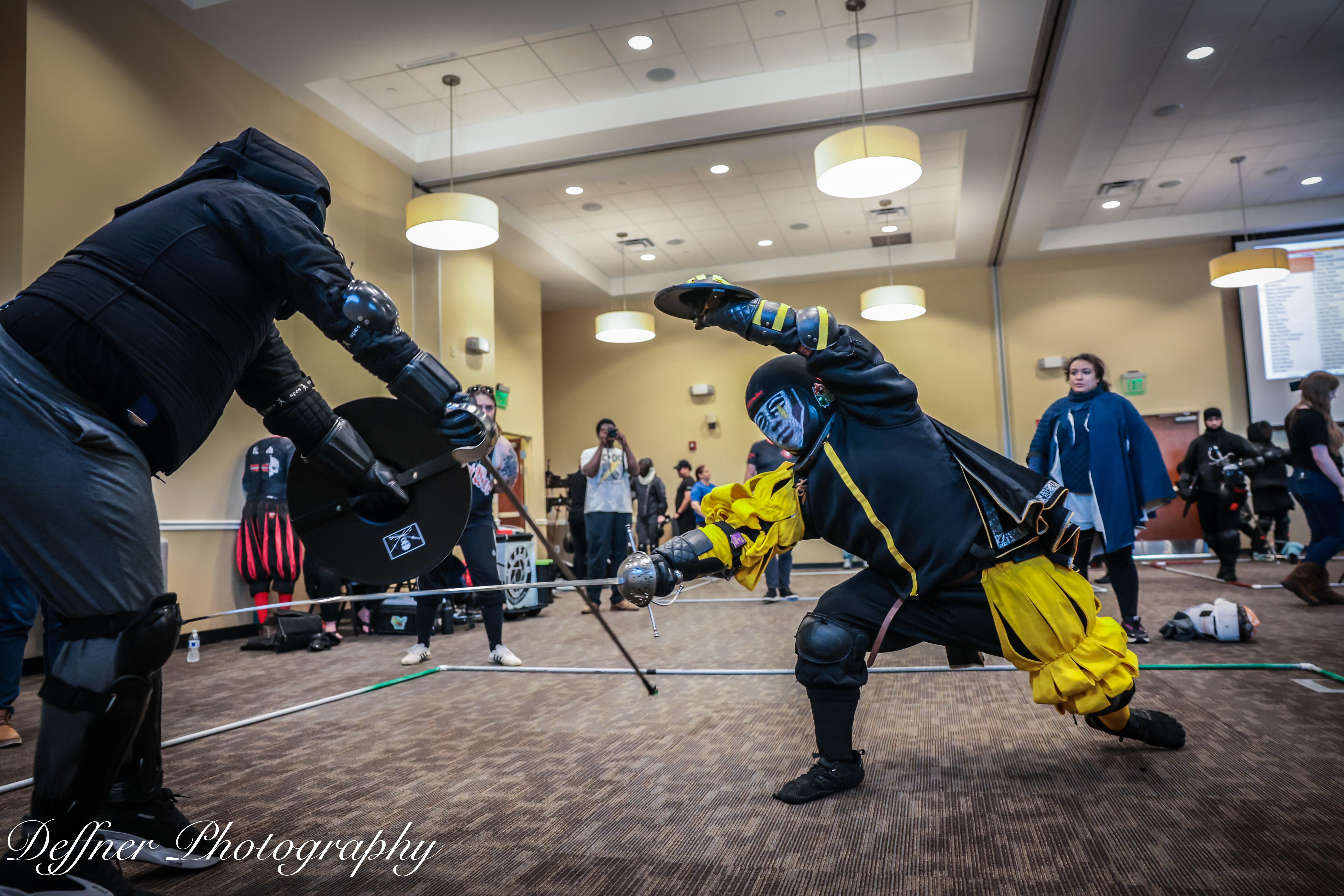 Two people in costumes perform a sword fight demonstration in a large indoor room with an audience watching.