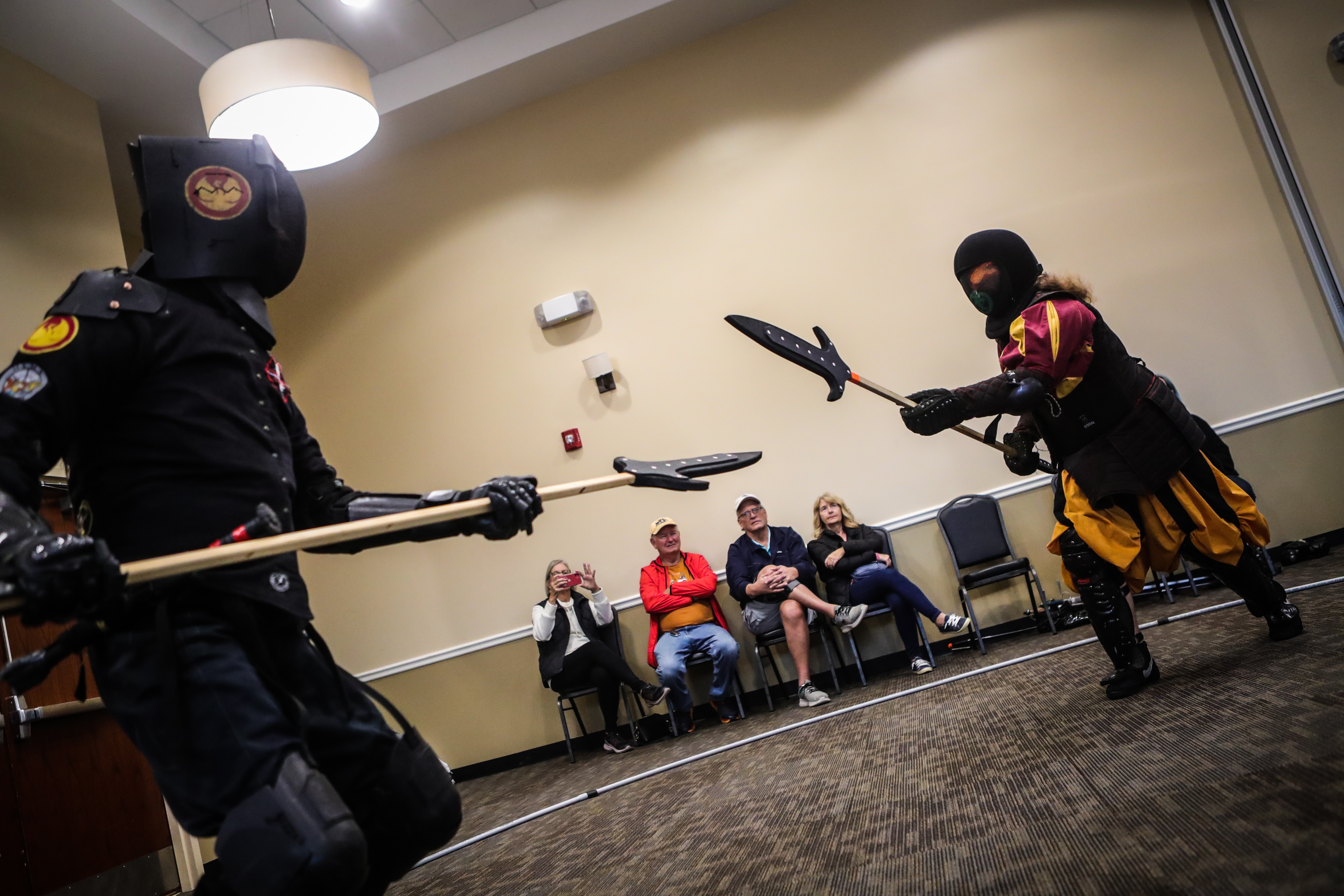 Two performers in costumes with weapons perform in front of seated audience members in a room.