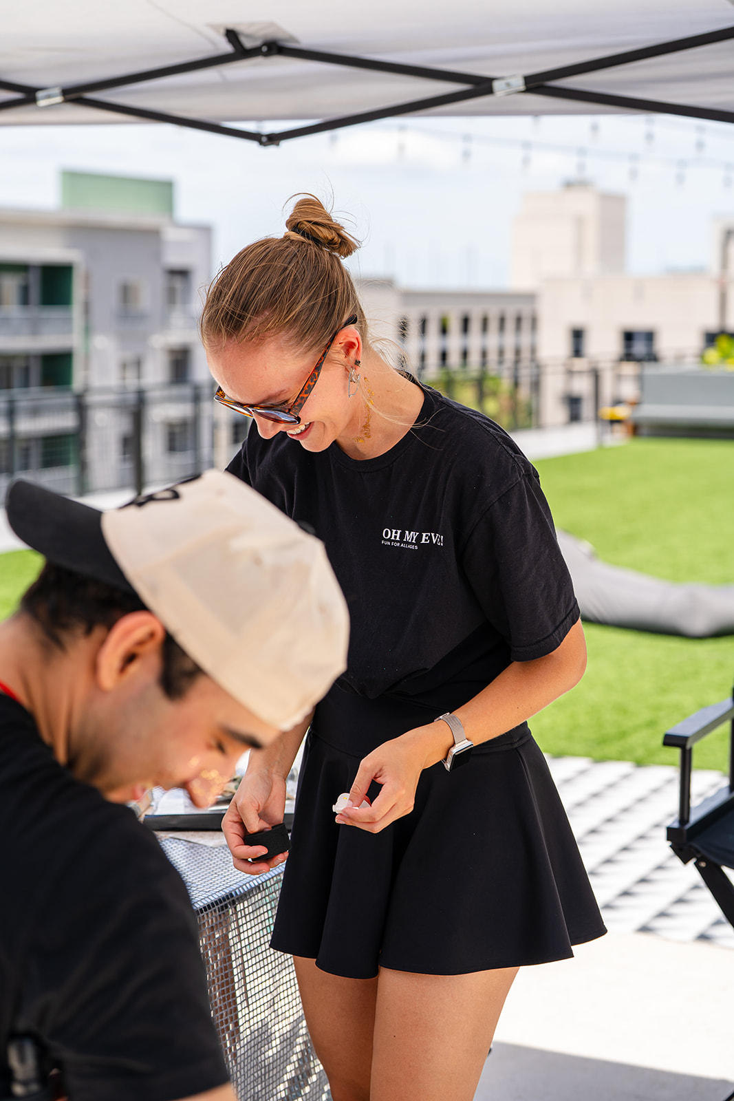 Two women under a canopy on a rooftop, one with a bun and glasses, the other wearing a cap, engaged in conversation.