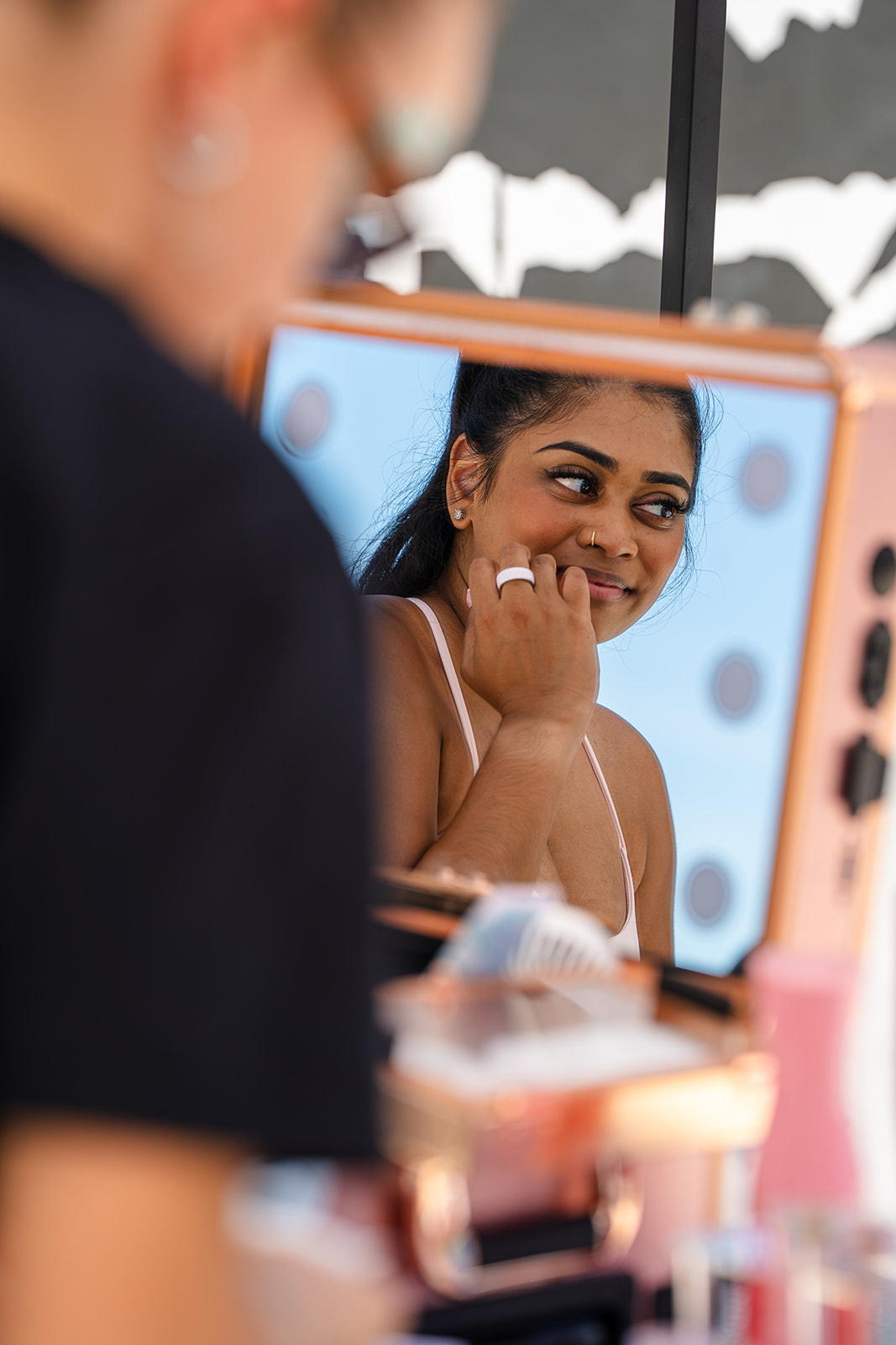 Young woman looking at herself in a mirror, smiling and touching her face, with a blurred person in foreground.