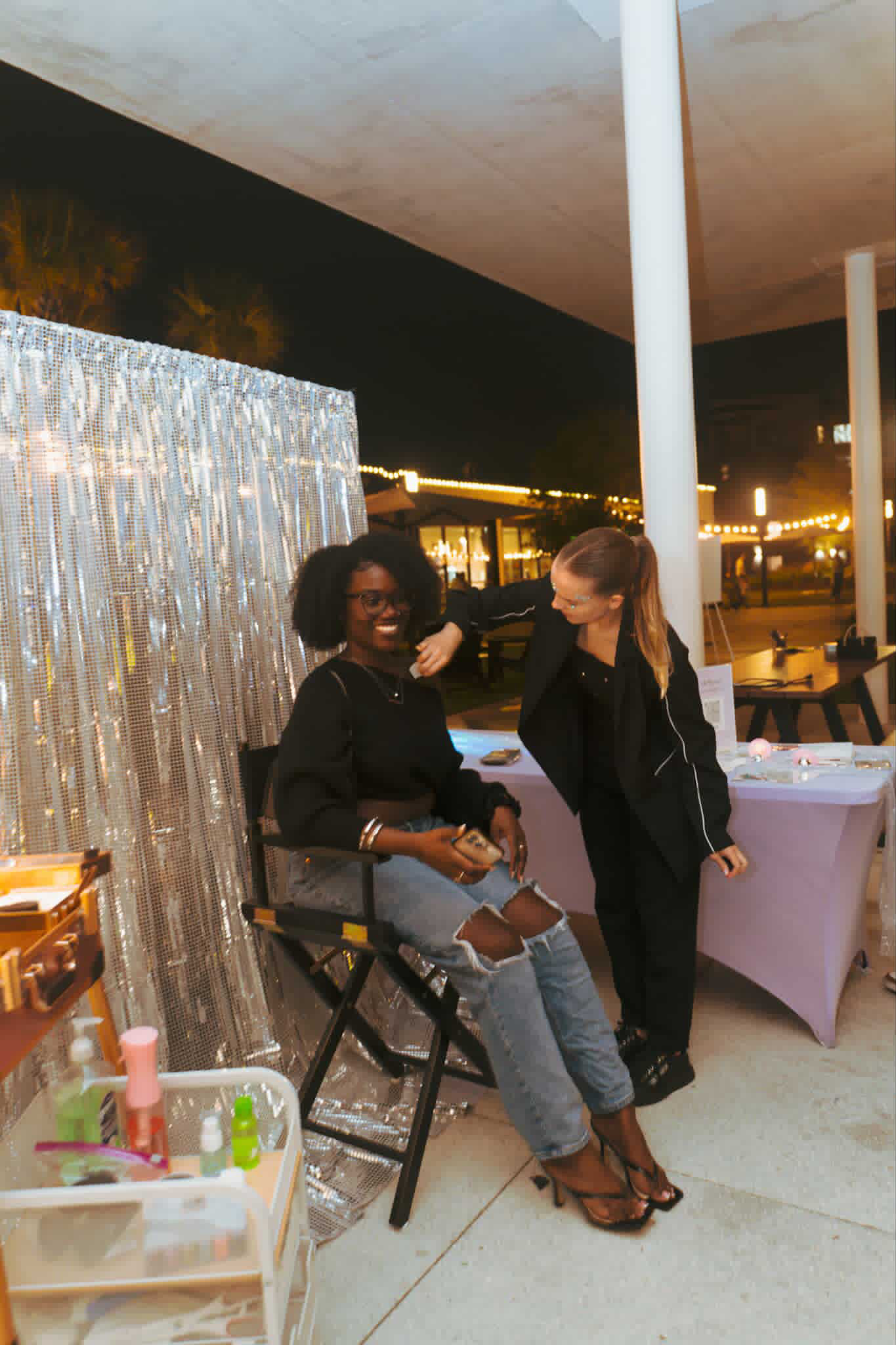 Two women at an outdoor event, one seated and the other standing, with a silver curtain backdrop and tables nearby.