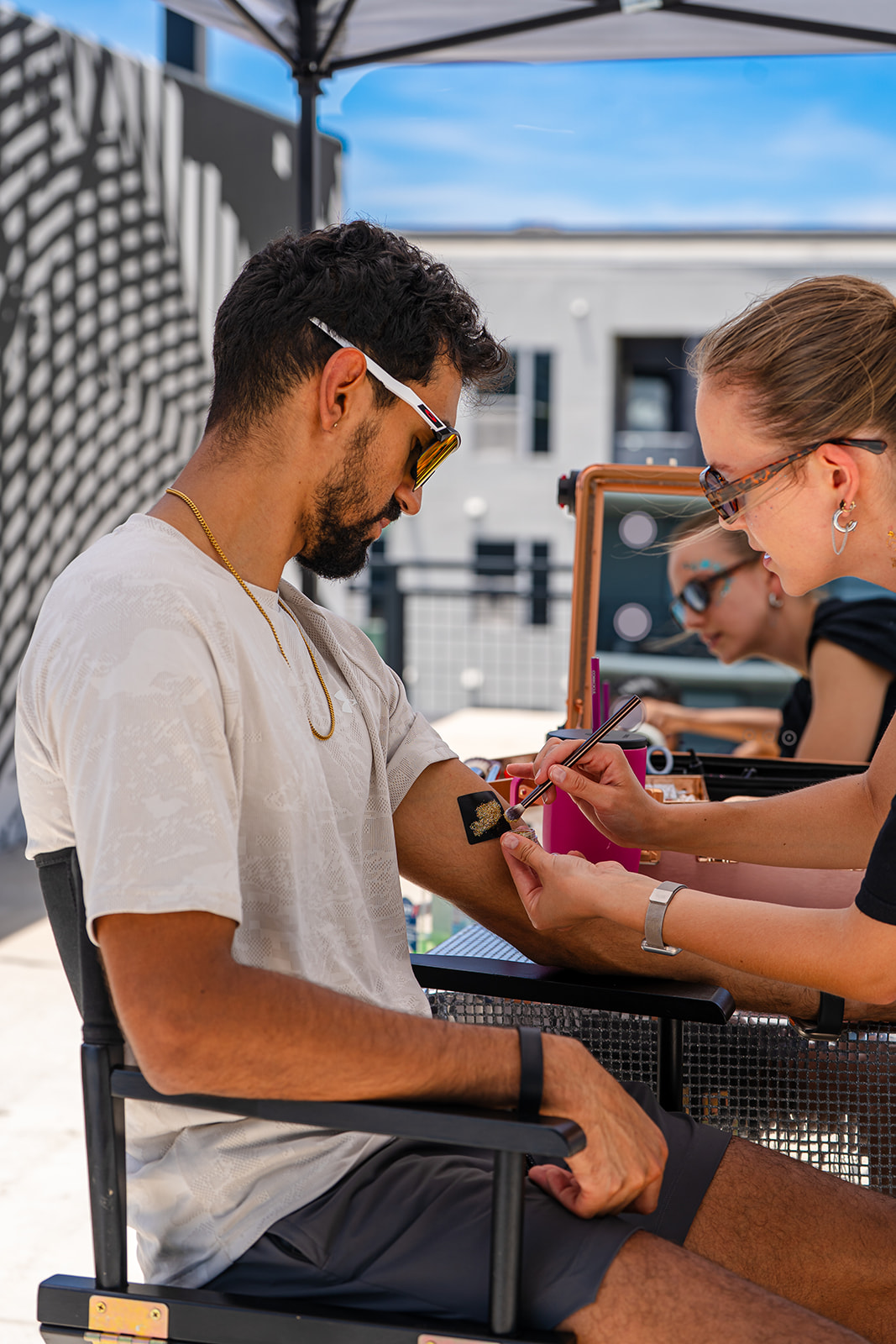 Man with sunglasses and a woman with glasses exchange a pink bag outdoors under a blue sky.