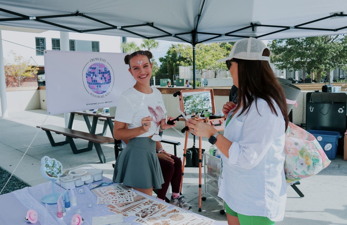 Two women talking under a canopy at an outdoor event, with tables and a whiteboard behind them.