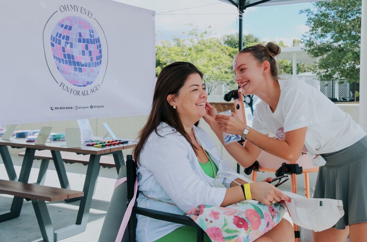 Two women smiling and talking outdoors, one sitting and the other leaning in, with a banner and tables in the background.