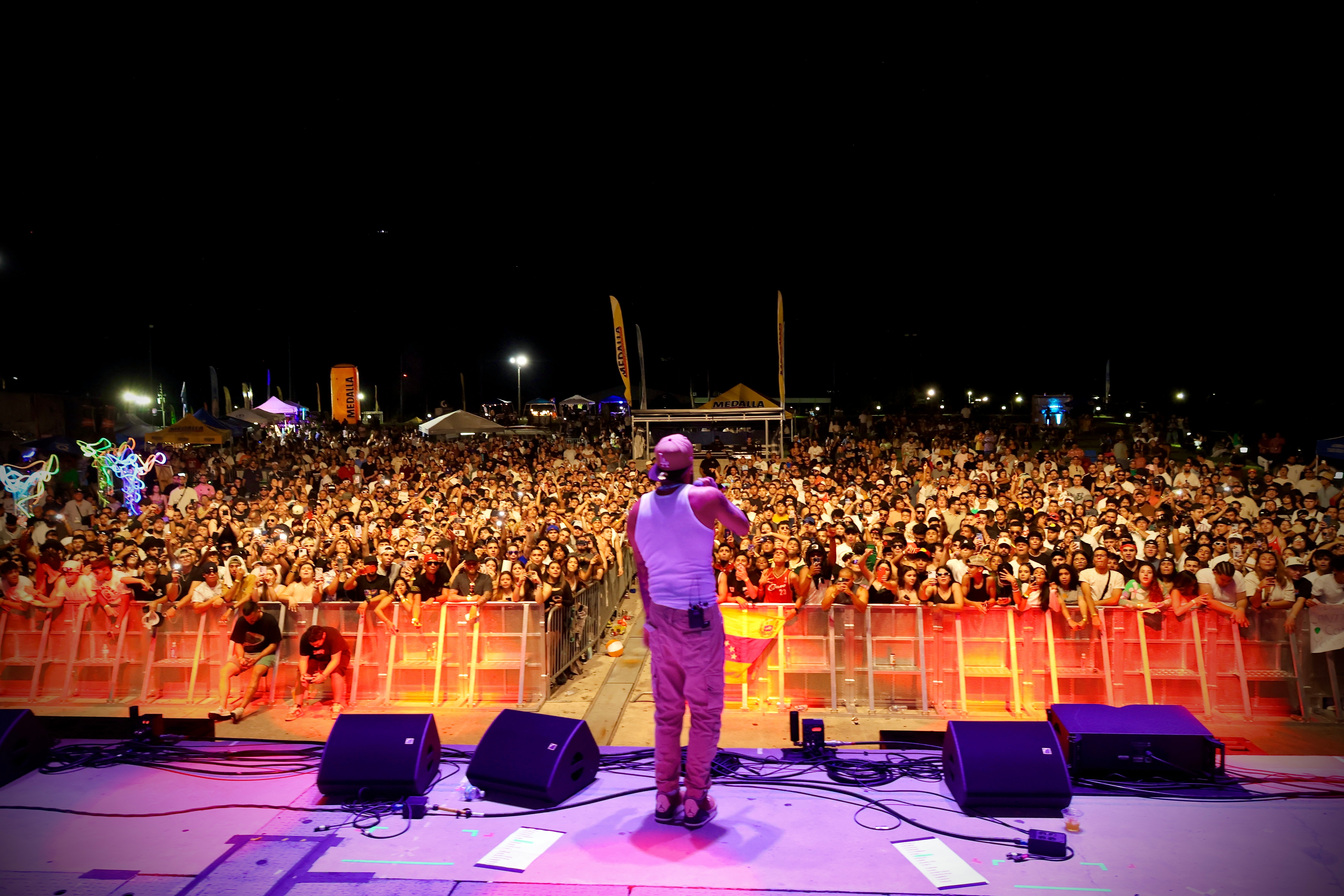 Performer on stage facing large crowd at night, stage lights illuminate scene, audience extends into distance, outdoor event setting.