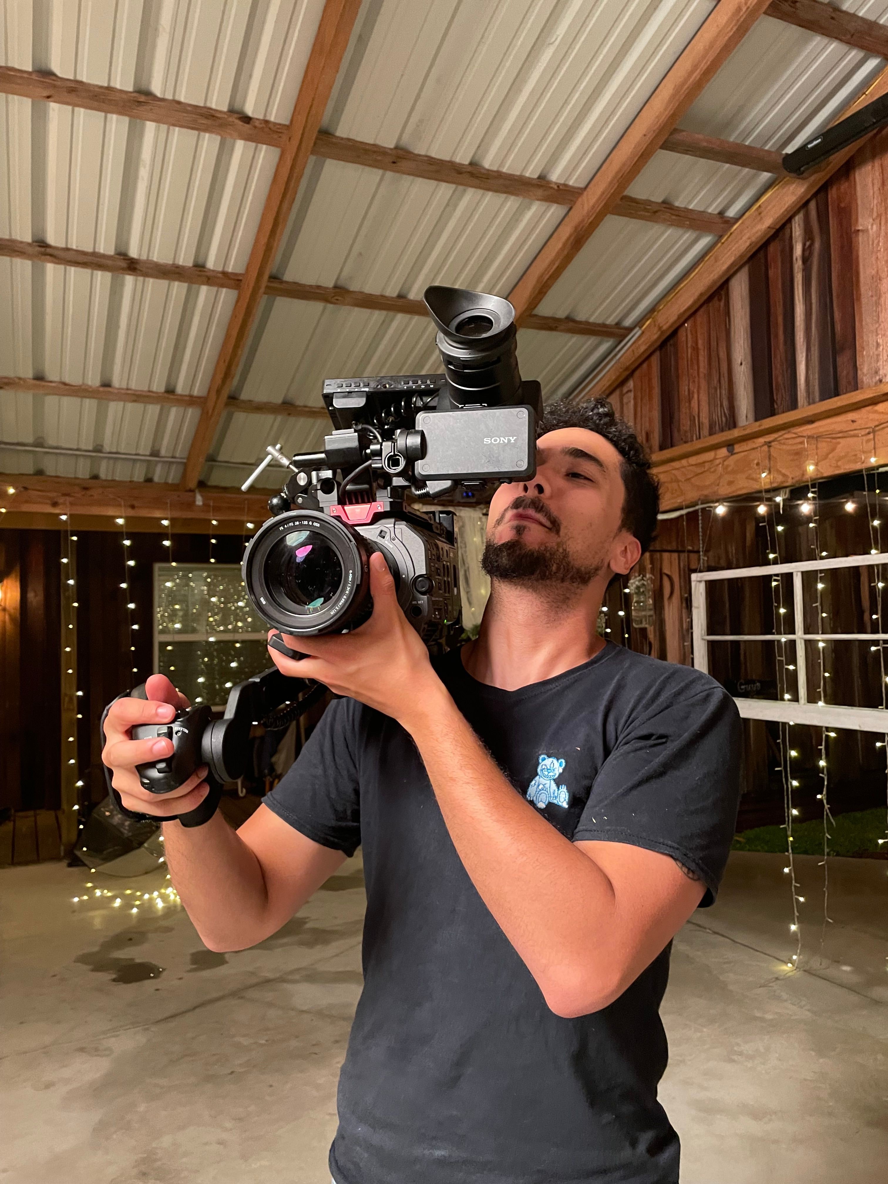 Person holding a camera with a stabilizer, smiling, in a wooden and string lights decorated space.