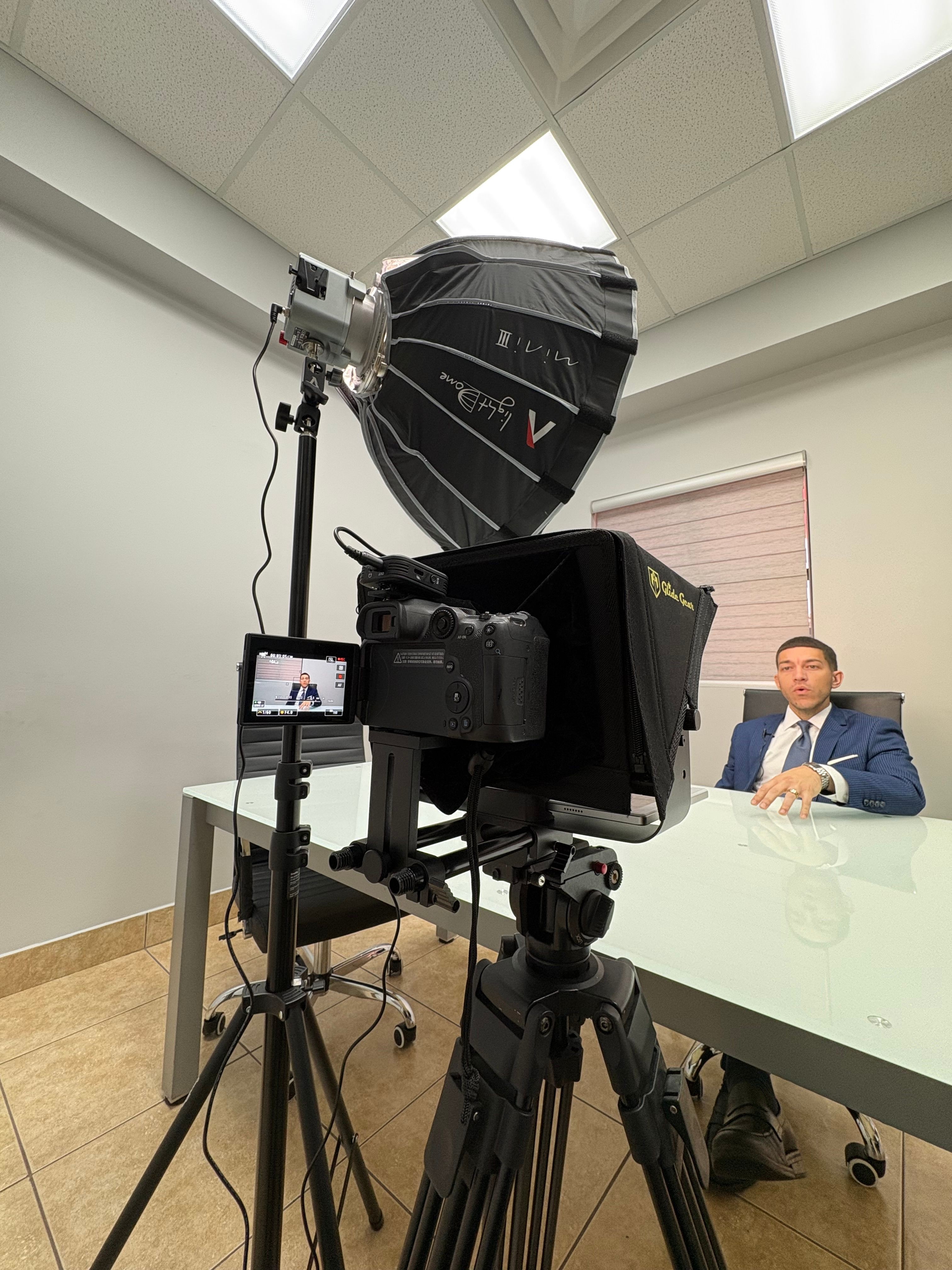 Person sitting at a table with a camera setup recording a man in a suit, in a room with white walls and ceiling.
