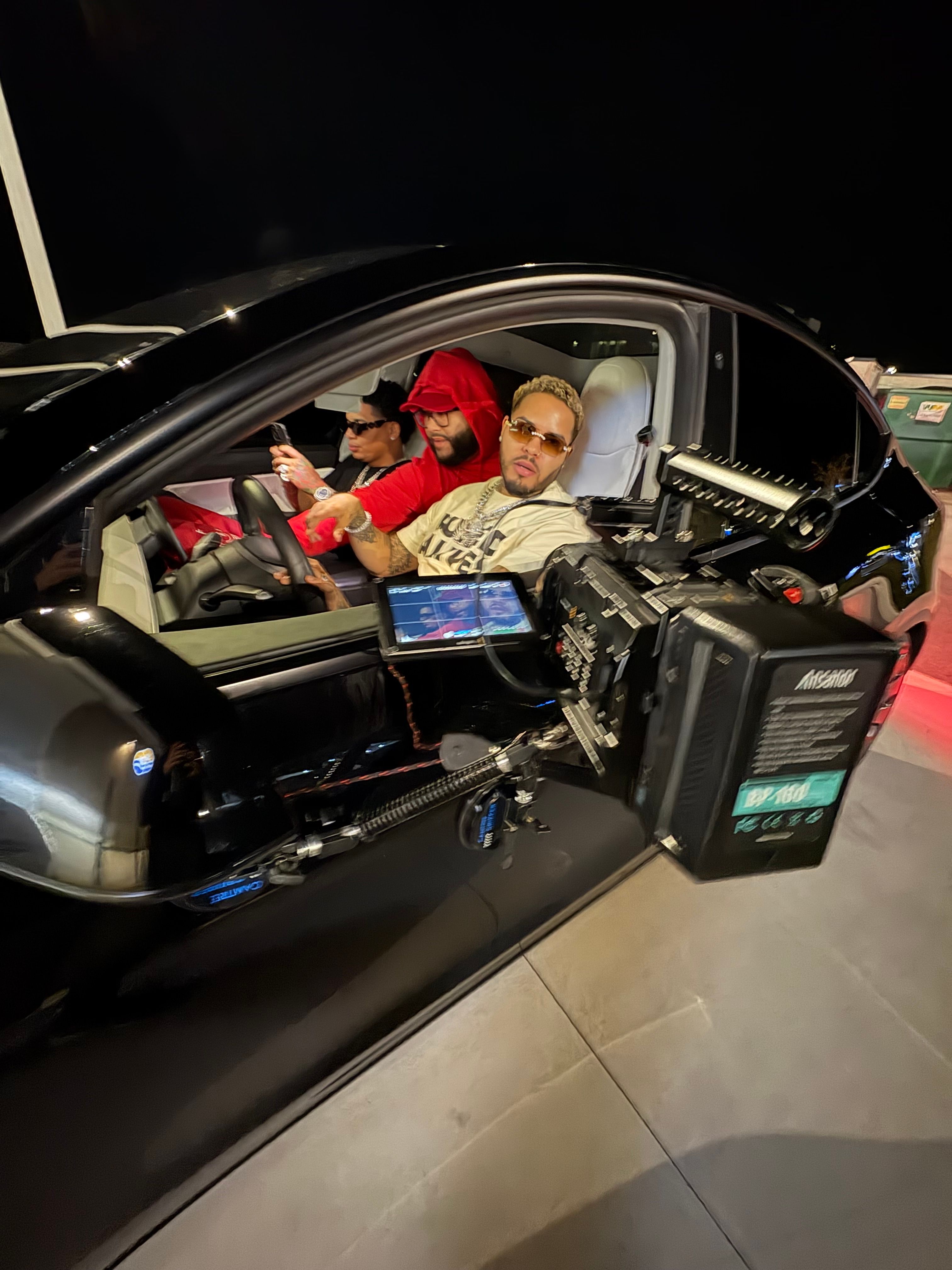 Two people sit in a vehicle with a camera and equipment mounted inside, looking up at the camera. The vehicle is indoors.