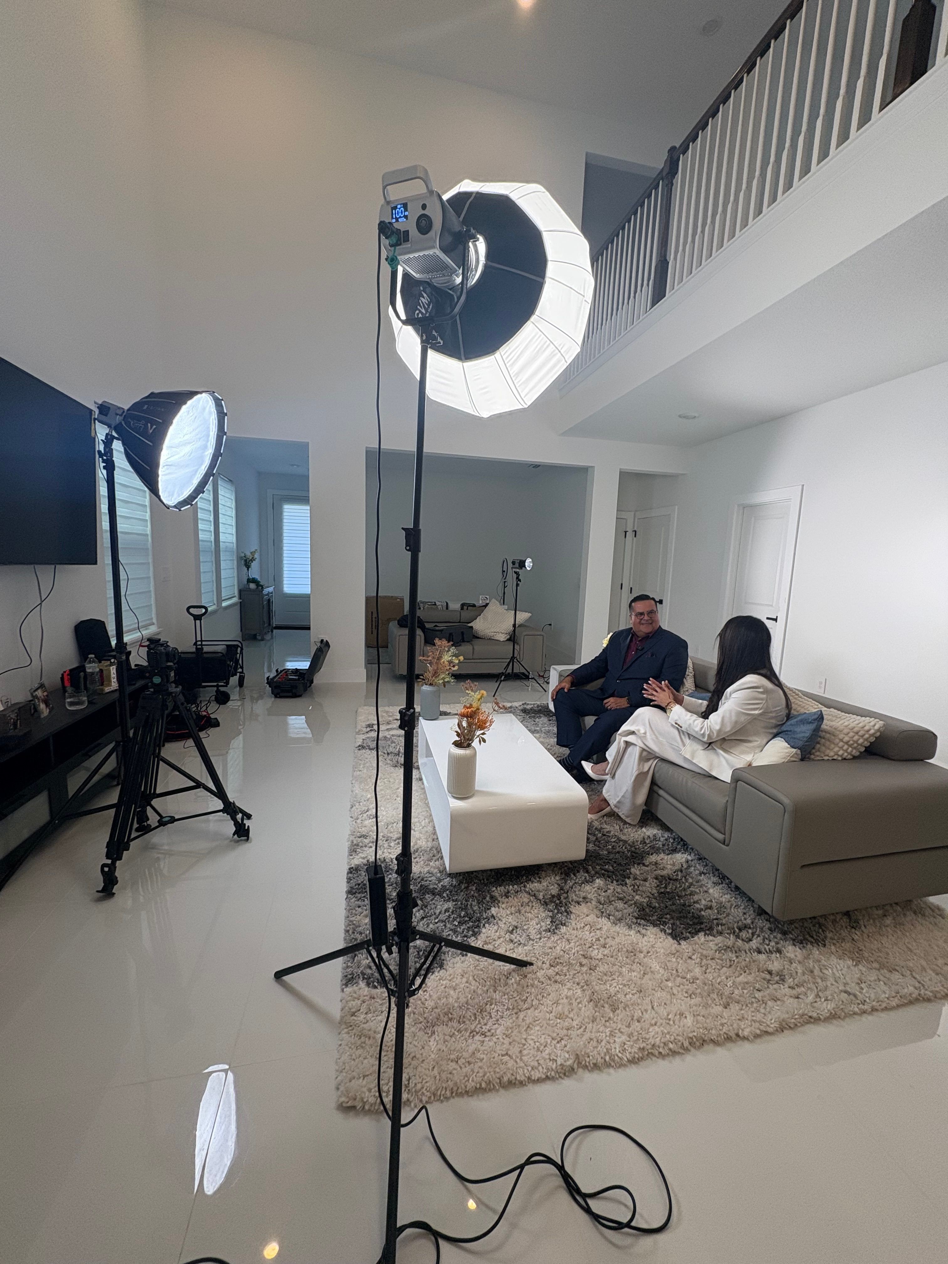 Two people sit on a sofa in a living room with a large ring light, camera equipment, and a tripod. Bright window light.