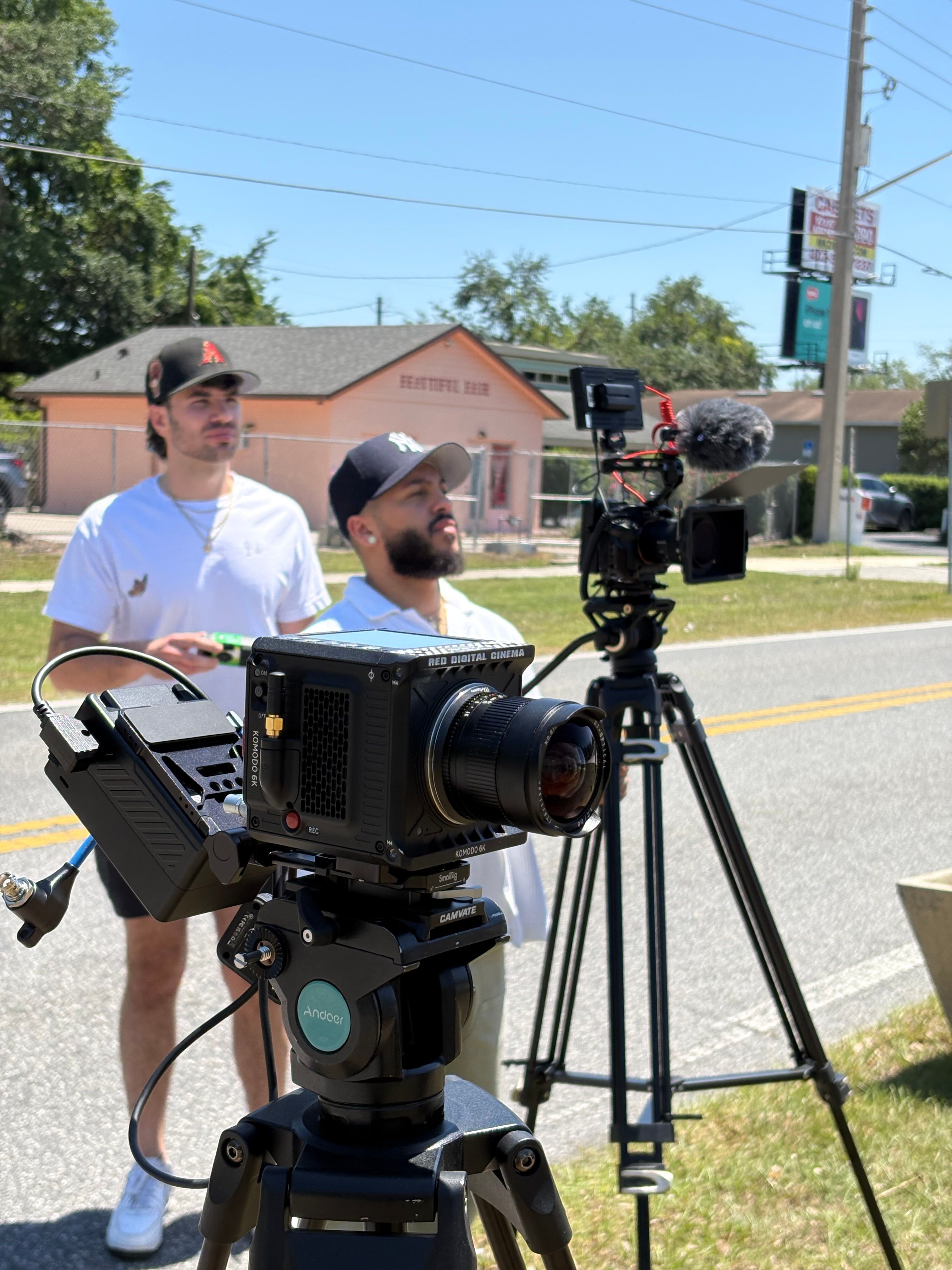 Two men stand outdoors near a camera setup, with a building and trees in the background.