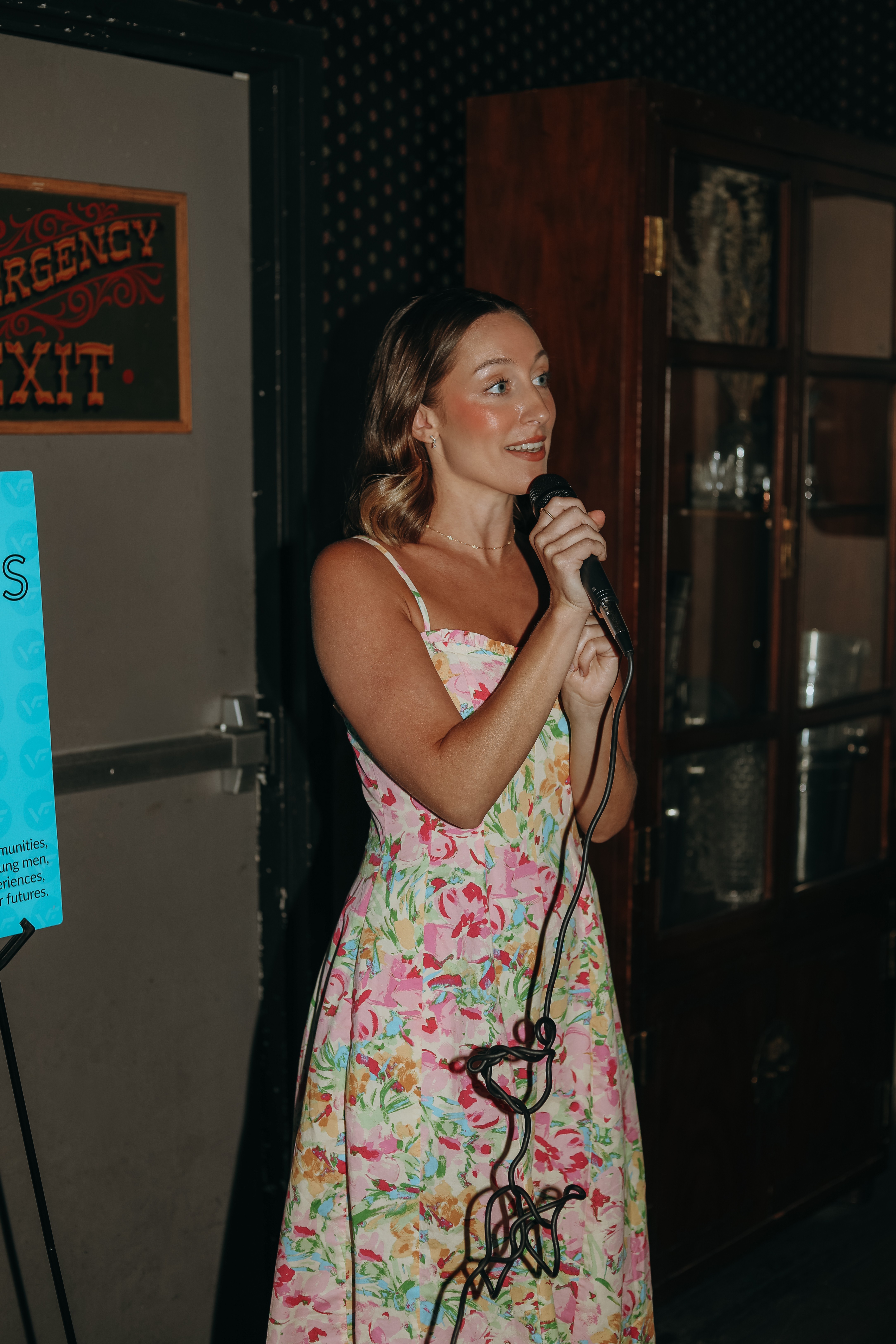 Woman holding microphone, smiling, in a floral dress, standing indoors near a door and window.