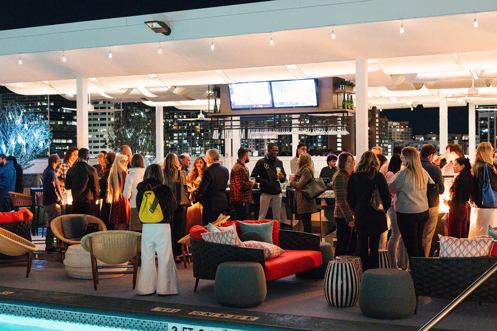 People gather in a modern indoor space with large windows, seating, and a bar area at night.