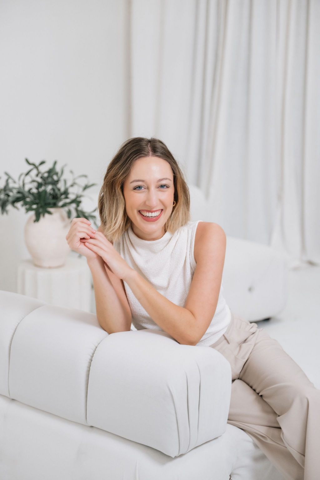 Smiling woman with shoulder-length hair sitting on a white sofa in a bright room with curtains and a potted plant.