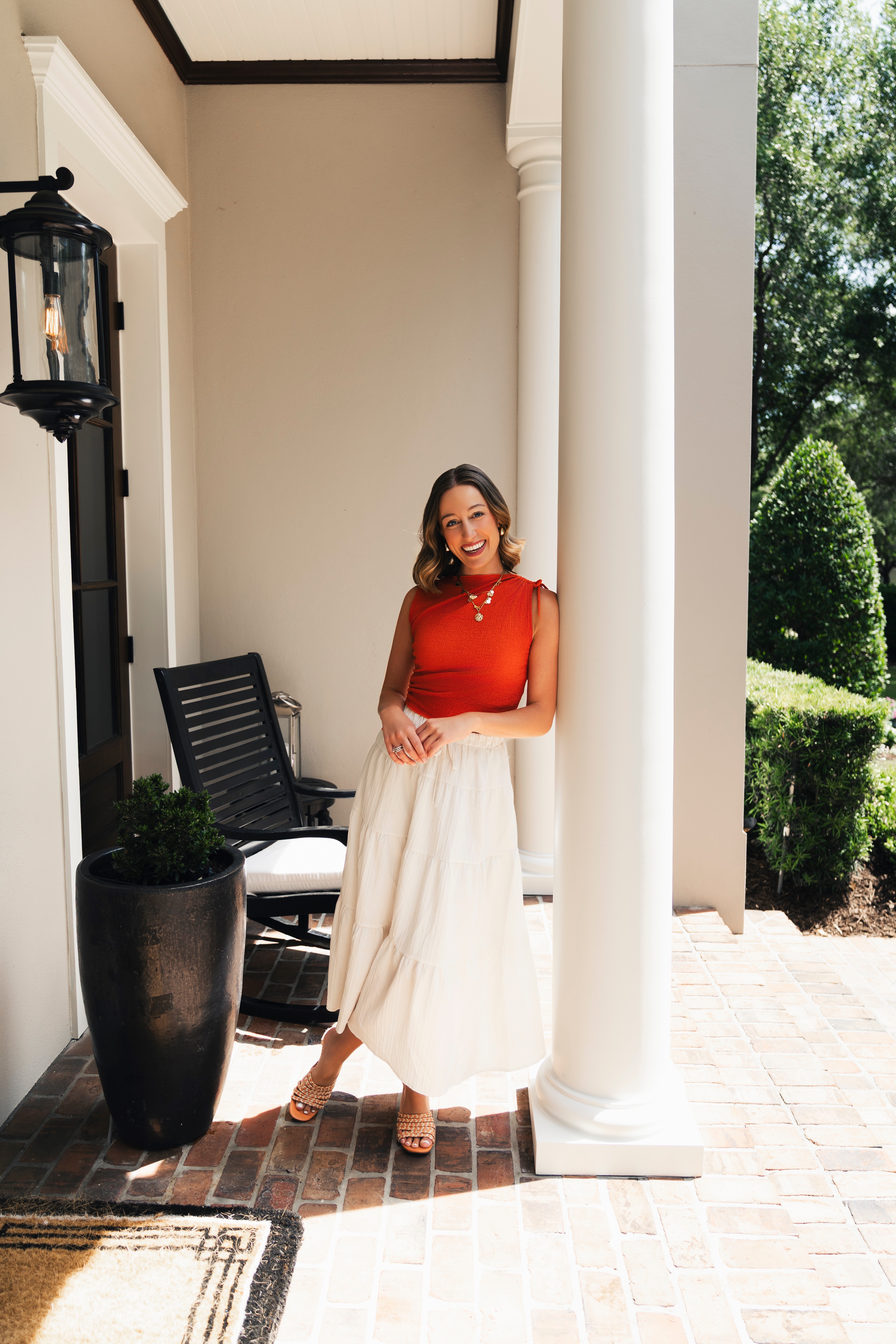 Woman in red top and white skirt leaning against a white column outside, with greenery in background.