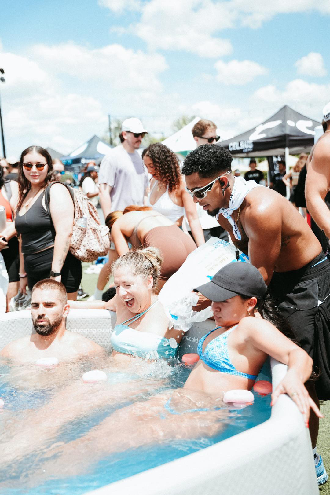 People gathered around a hot tub outdoors, some in swimsuits, with tents and a blue sky in the background.