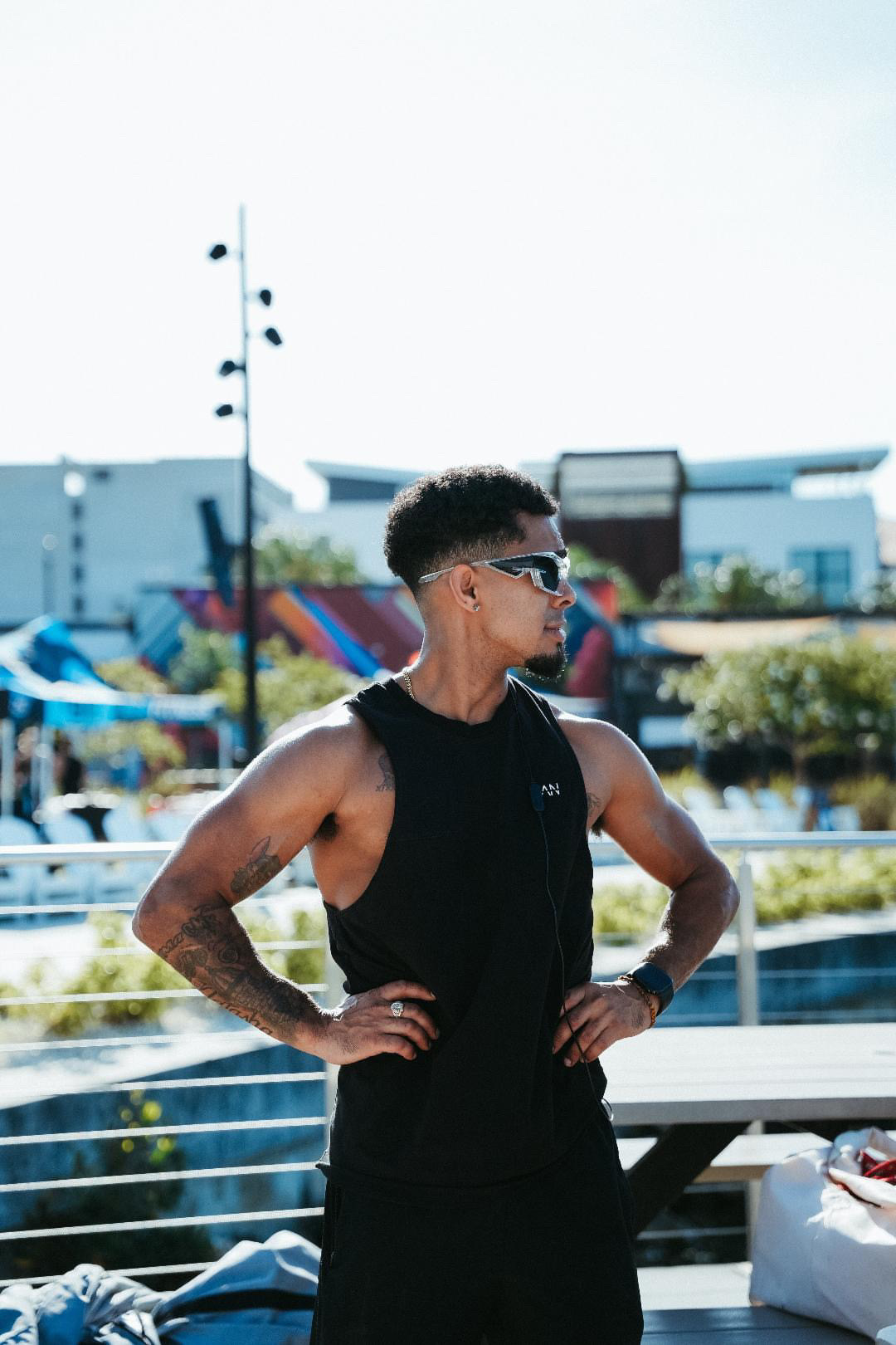 Young man with sunglasses and tattoos stands outdoors with hands on hips, wearing a black sleeveless shirt.