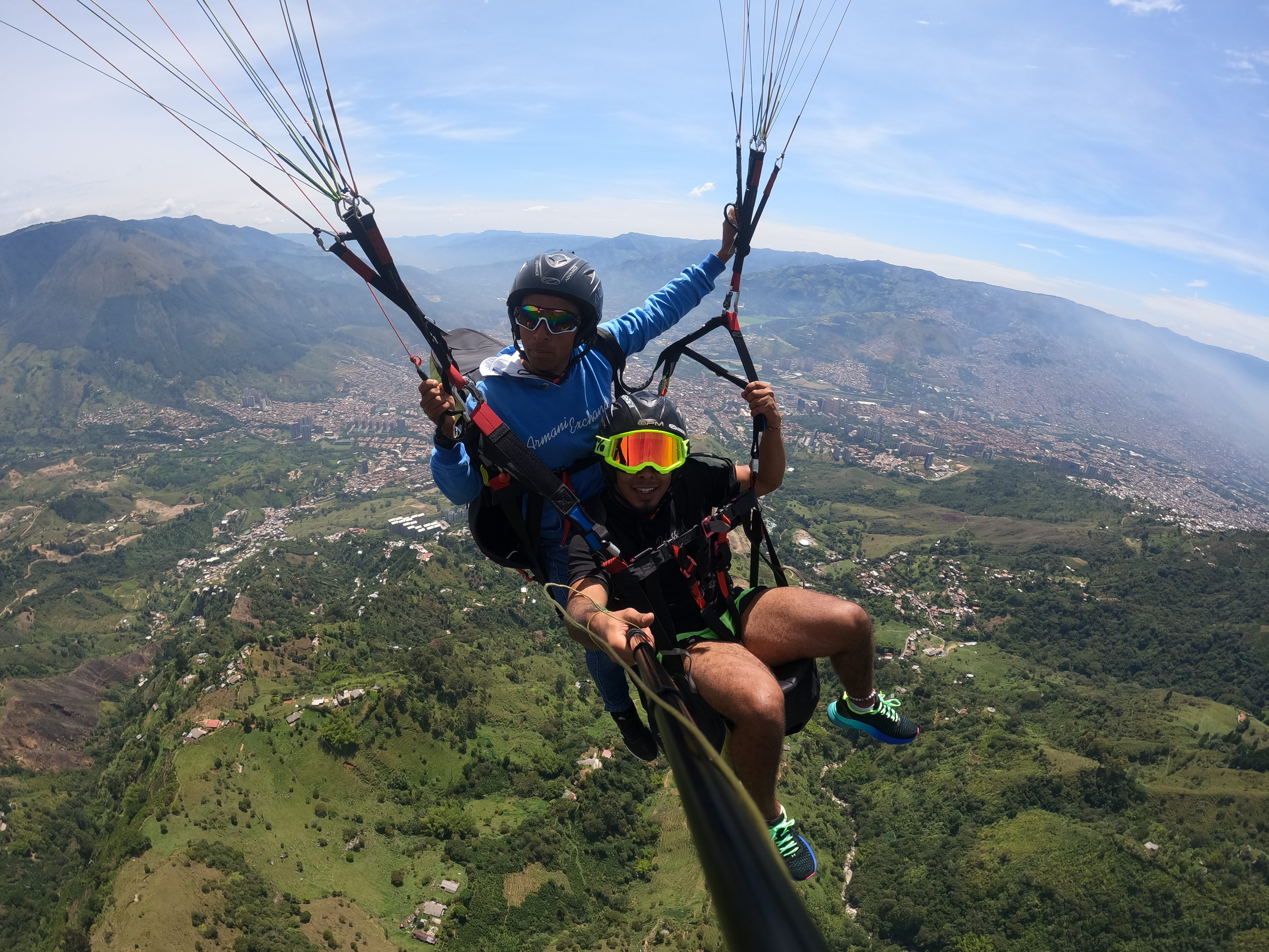 Person paragliding high above green landscape with mountains and a town below, wearing helmet and goggles.