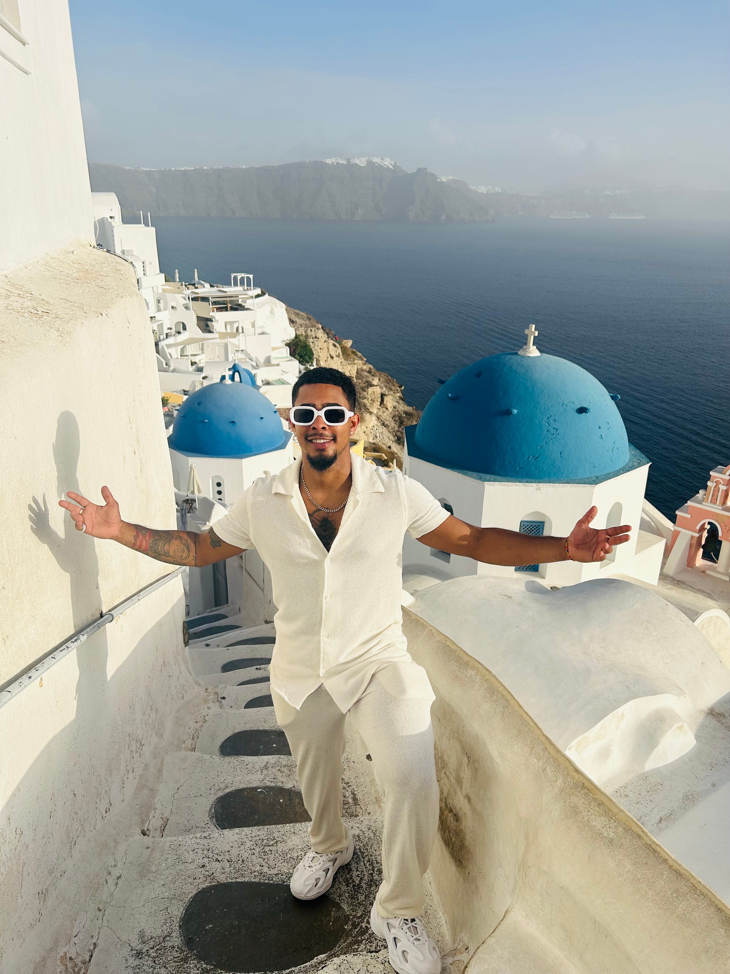 Man in white outfit and sunglasses standing on a white rooftop with blue domes overlooking the sea and cliffs.