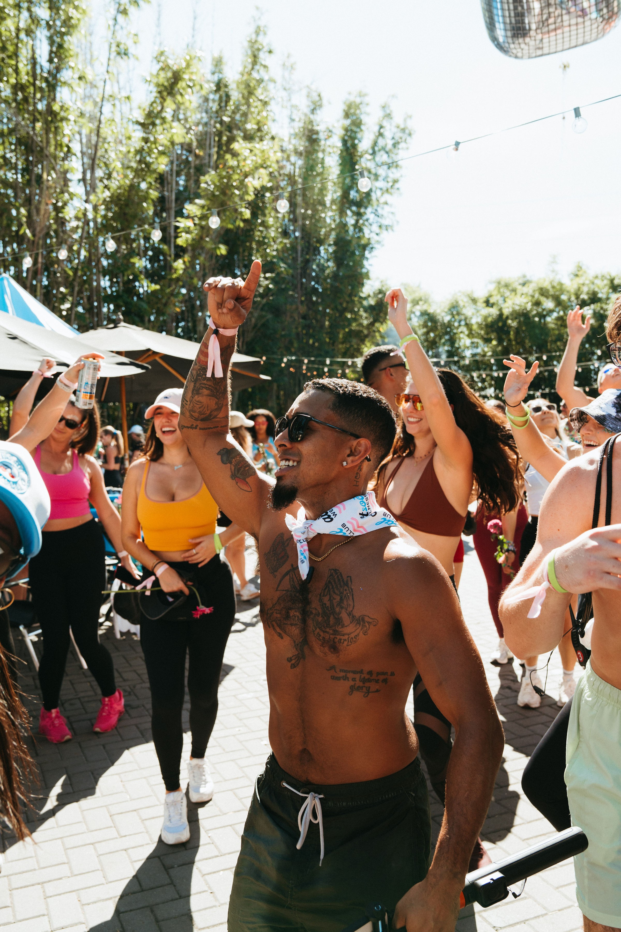 Group of people dancing outdoors with trees and string lights in background, sunny day, diverse group, lively atmosphere.