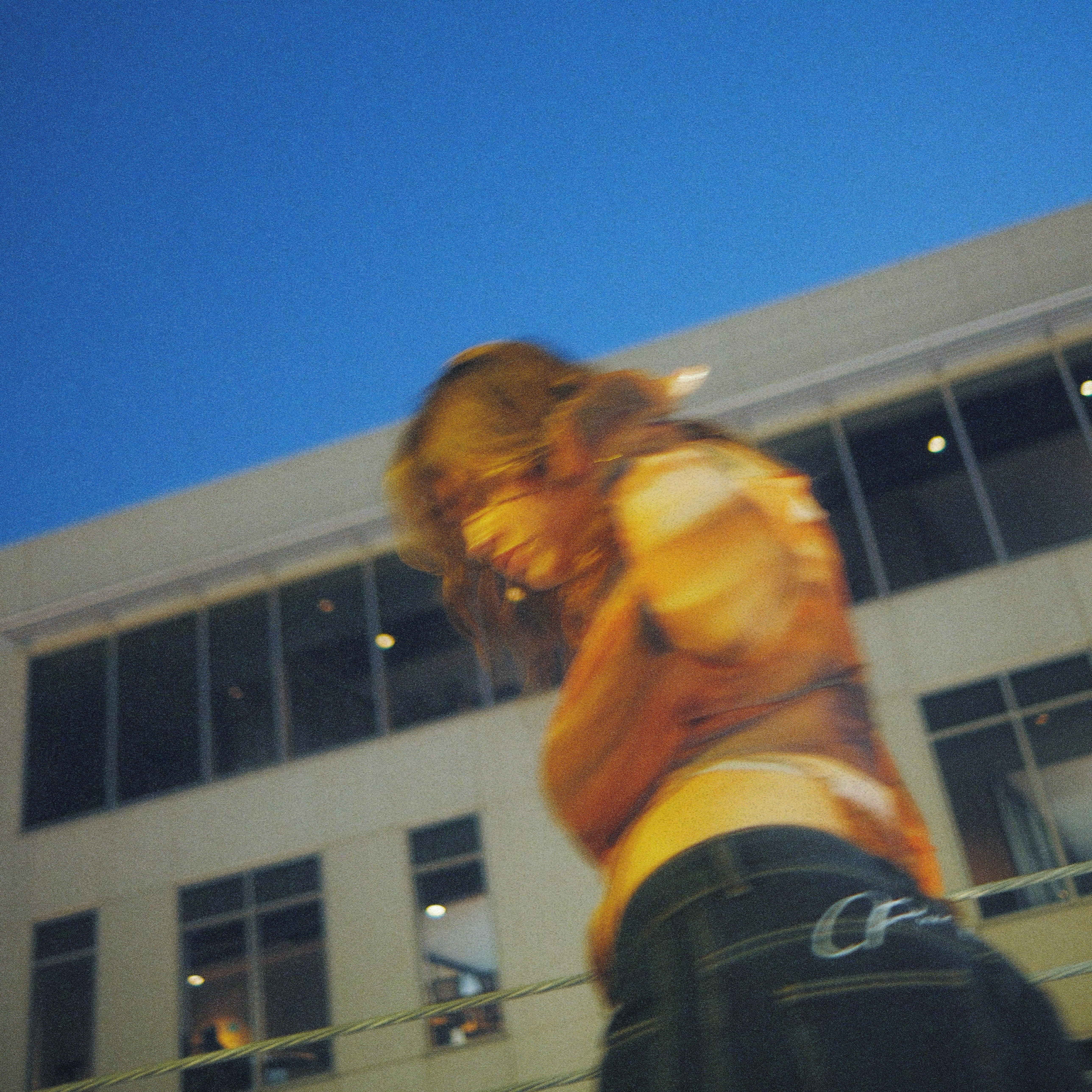 Person with long hair wearing a yellow top and dark jeans, standing outside near a modern building, looking down.