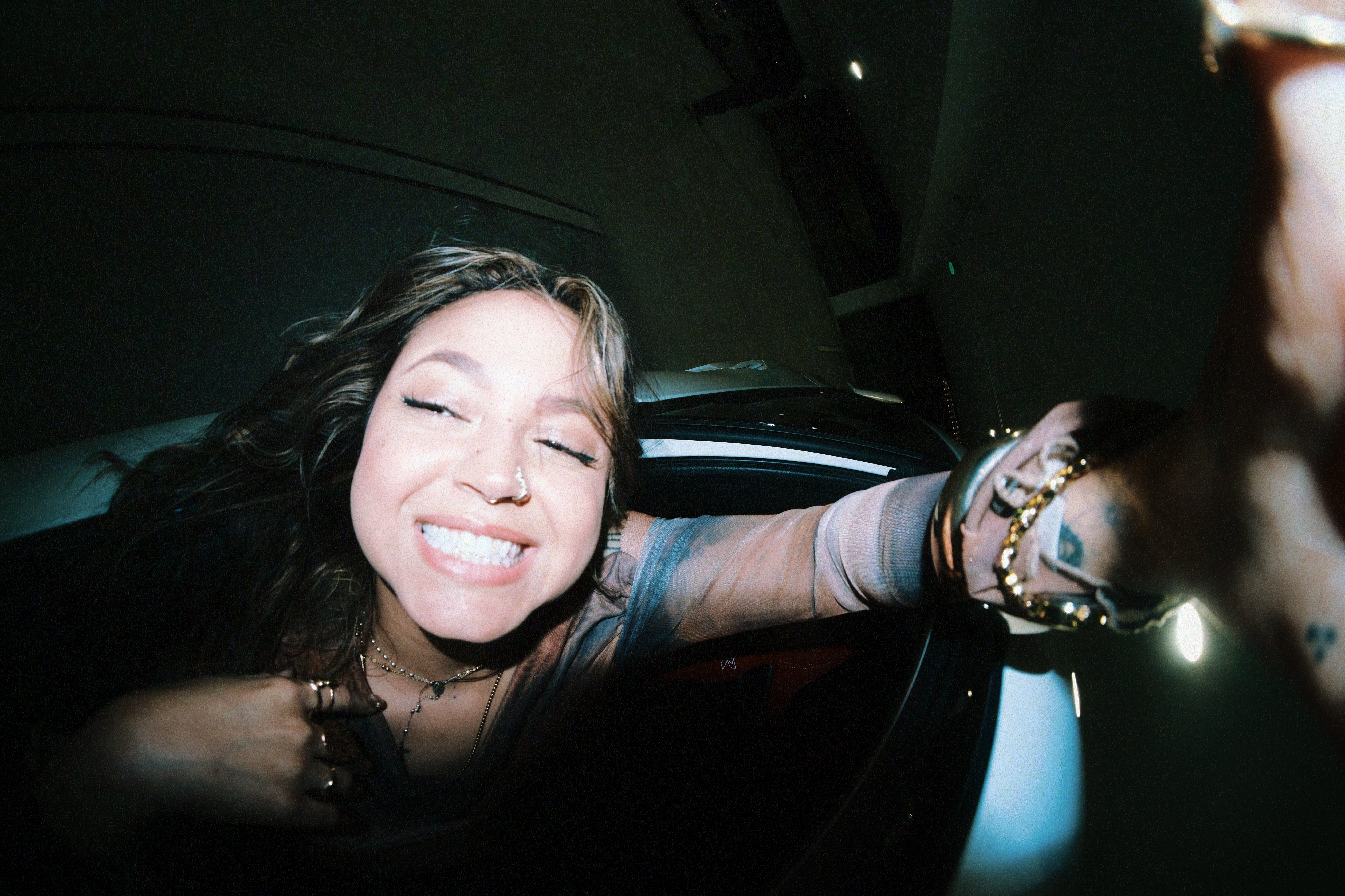 Woman smiling with tongue out, taking selfie inside a car at night, wearing a striped shirt and jewelry.