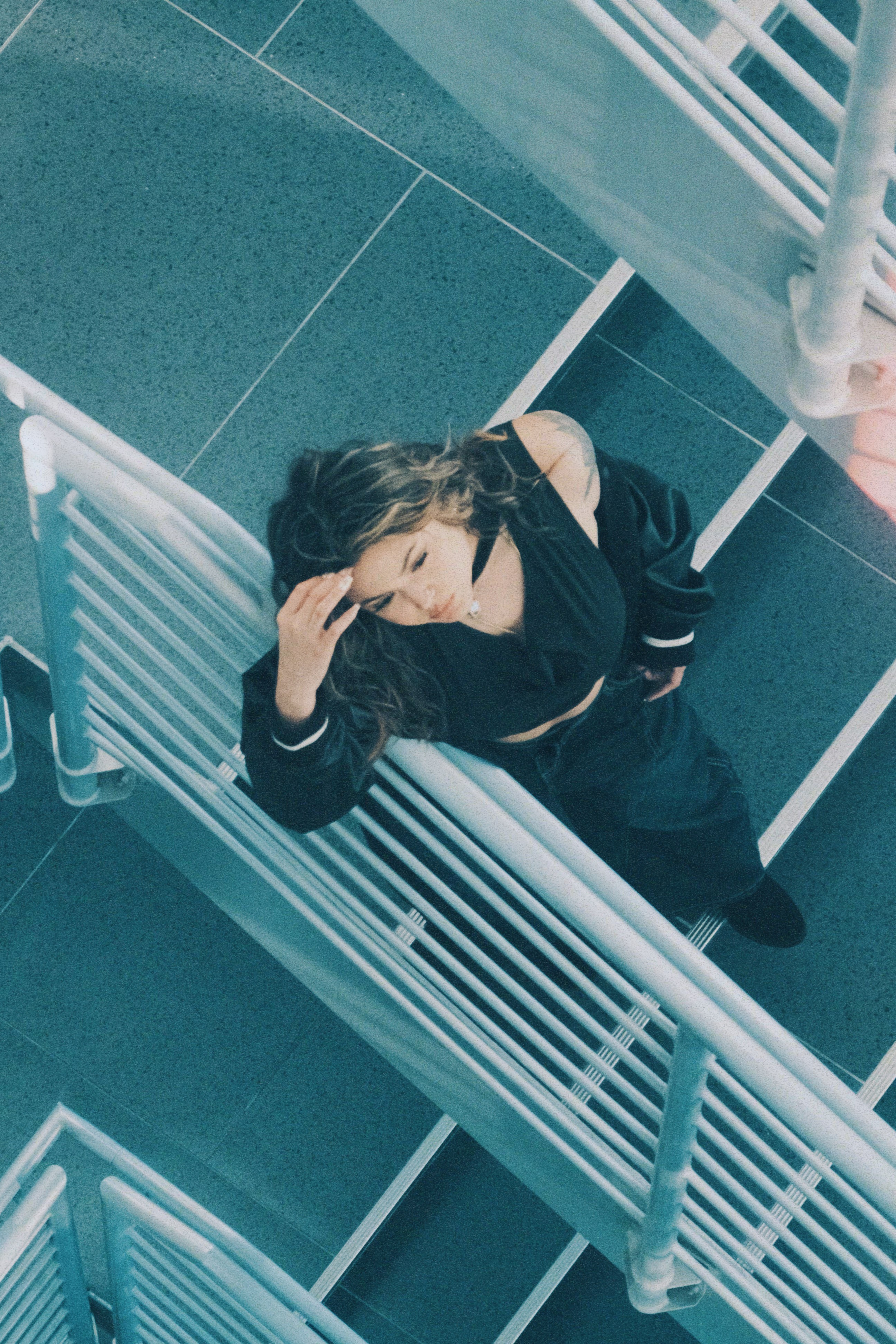 Woman with dark hair in black outfit standing on a white staircase, looking up, with teal tiled floor below.