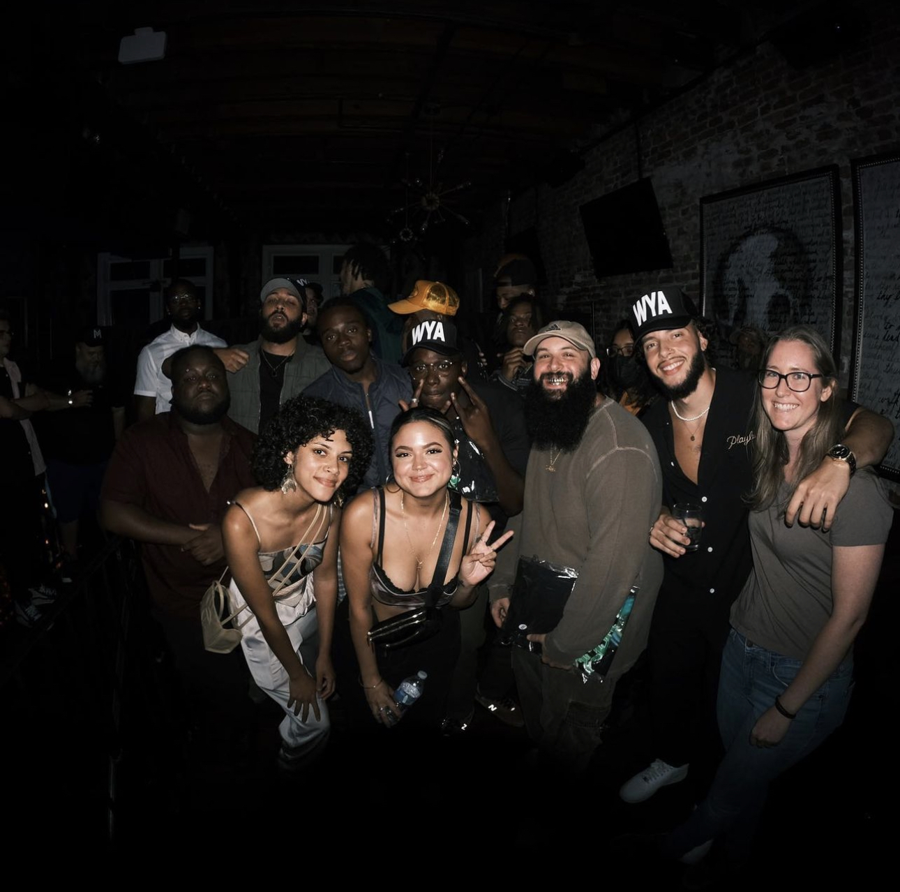 Group of people smiling and posing together in a dimly lit indoor setting, some wearing hats with 'WYA' written on them.